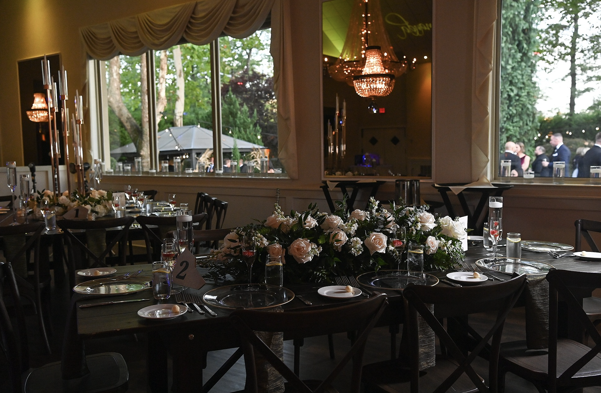 Elegant event table with flower centerpiece, place settings, and candles in a decorated room with large windows showing an outdoor patio area.