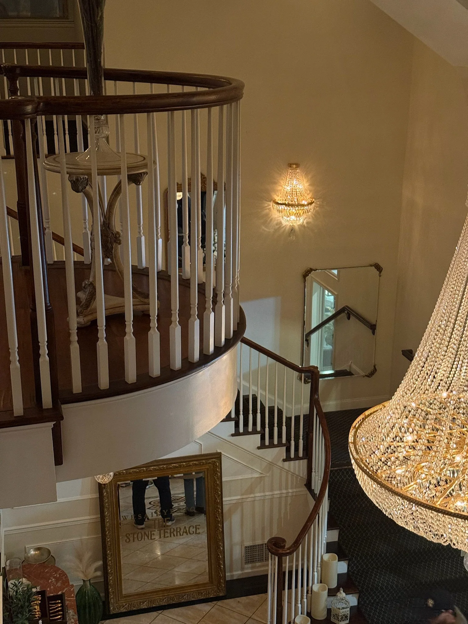 View of an elegant staircase with dark wood handrails and white balusters, a large ornate chandelier hanging above, and a mirror reflecting the staircase. The setting appears to be in a luxurious home or hotel foyer.