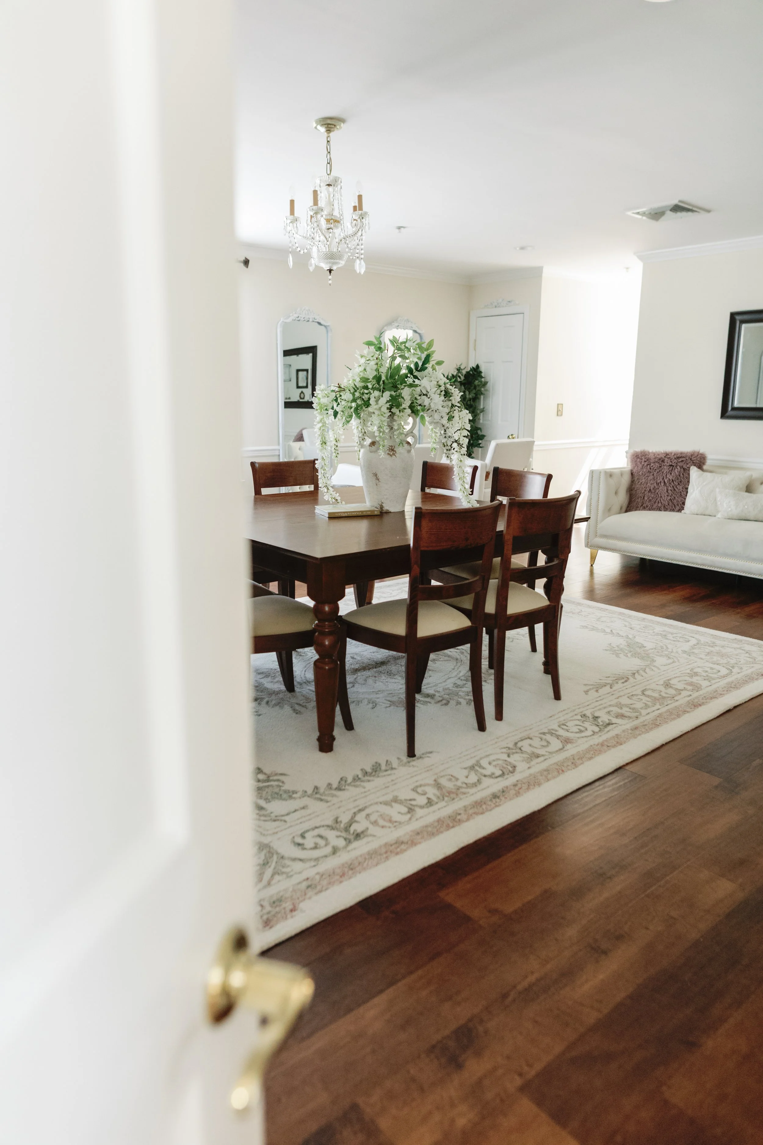 View of a dining room with a wooden table, six chairs, a large white vase with white flowers, and a chandelier hanging from the ceiling.