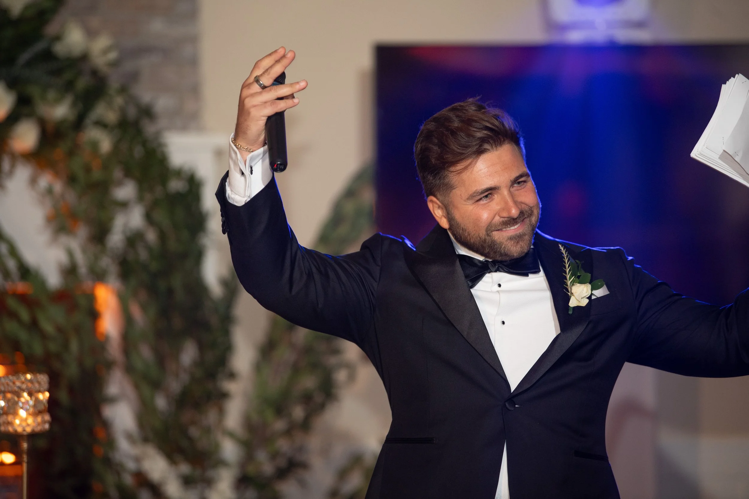 Man in tuxedo smiling and waving at a wedding or formal event.
