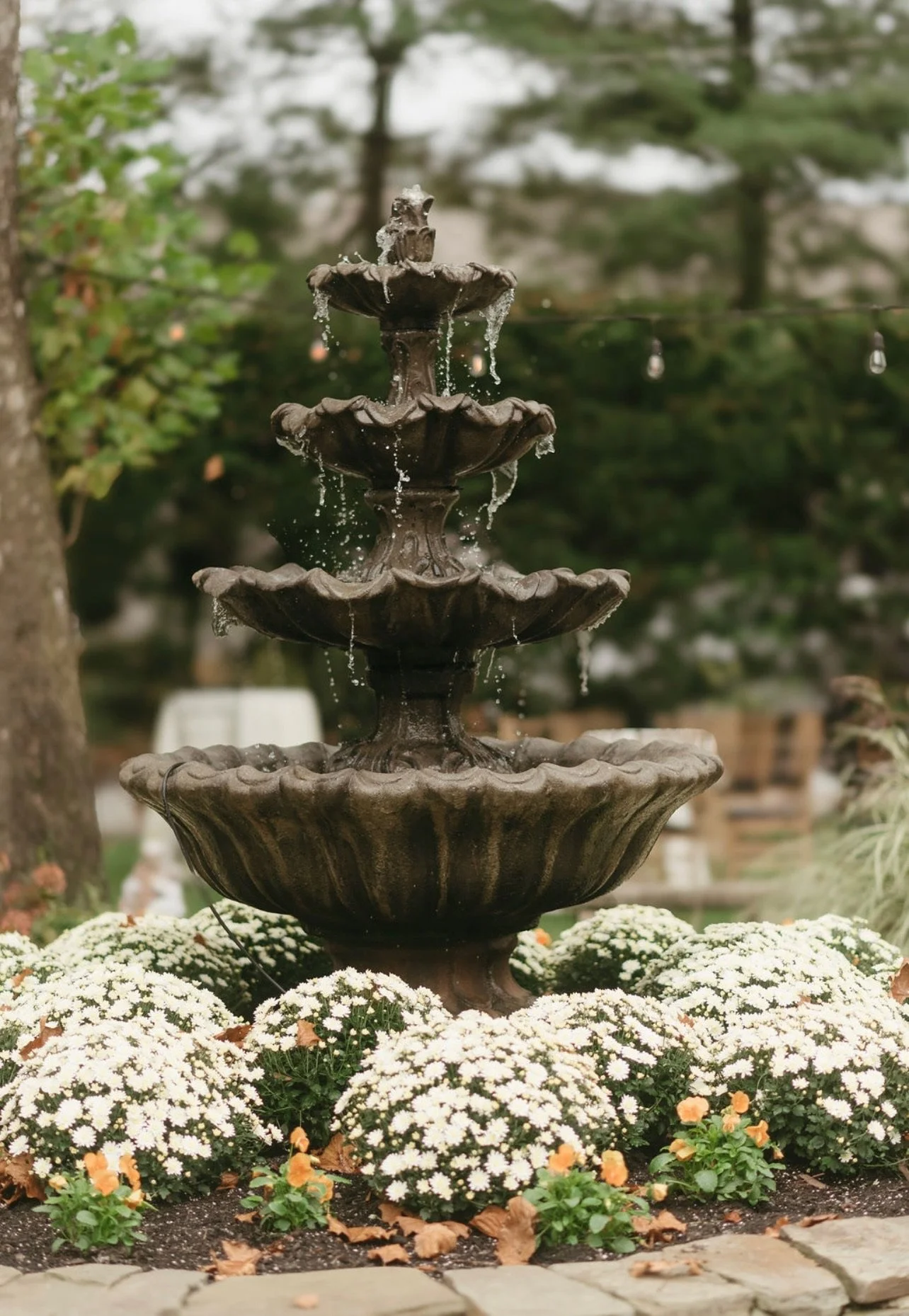 A multi-tiered outdoor fountain surrounded by white and orange flowers, set in a garden with trees in the background.