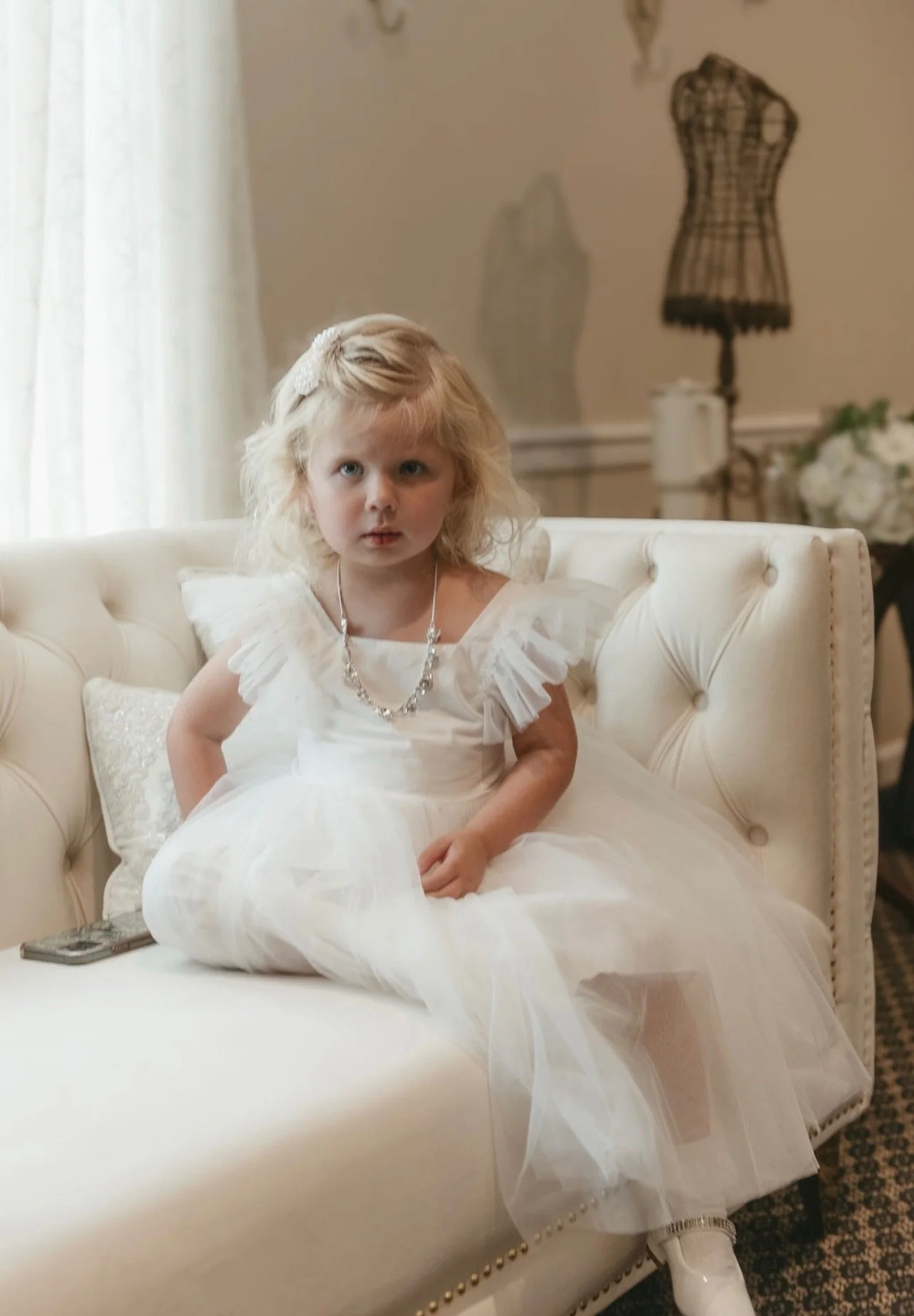 A young girl in a white dress sitting on a cream-colored sofa in a room with soft lighting.
