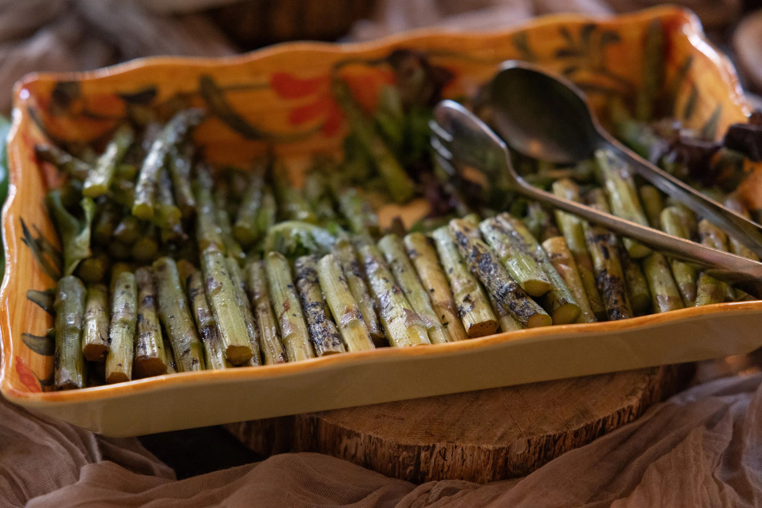 A ceramic baking dish filled with roasted asparagus spears on a wooden cutting board with serving tongs.