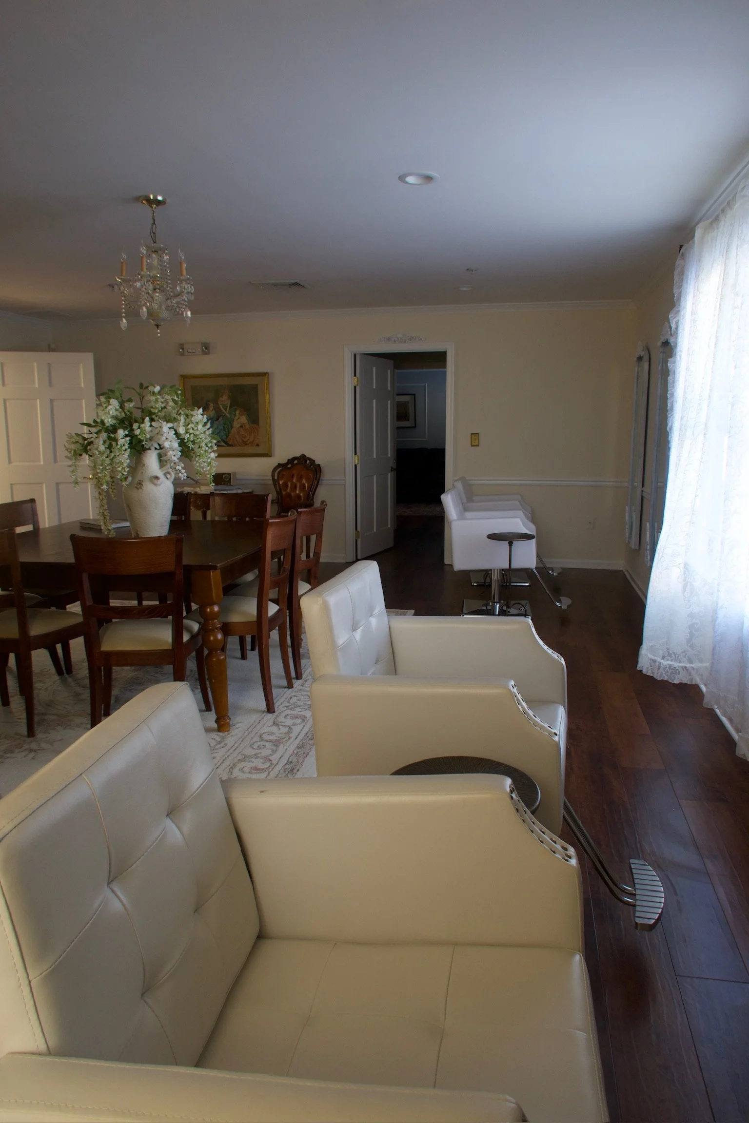 Interior of a living room with beige chairs, a wooden dining table with a floral centerpiece, a chandelier, and a doorway leading to another room with white barstools along a counter.