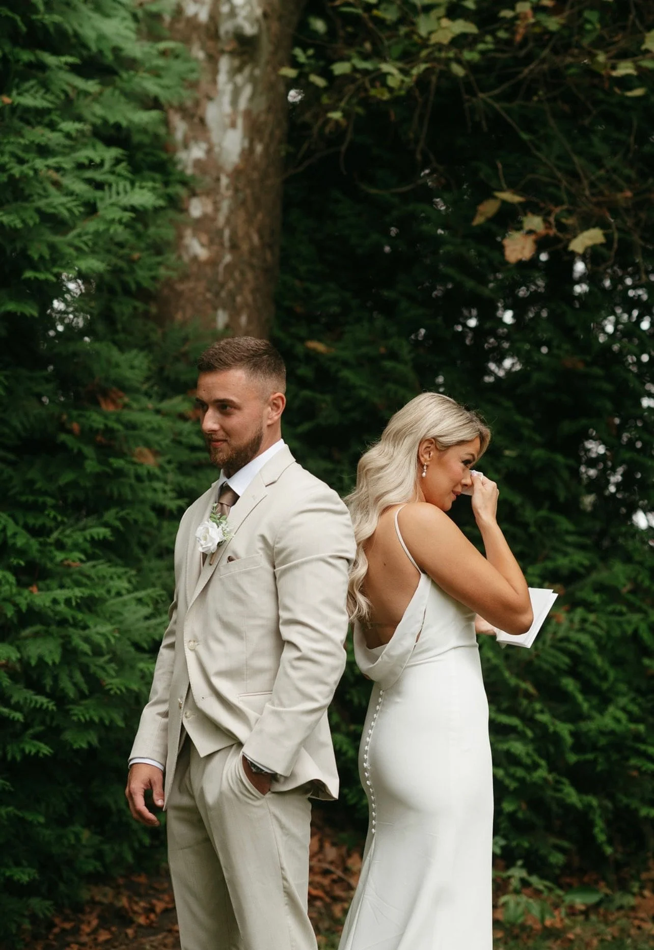 A newlywed couple standing outdoors among green trees, with the bride wiping away tears and the groom standing with hands in pockets, both dressed in wedding attire.