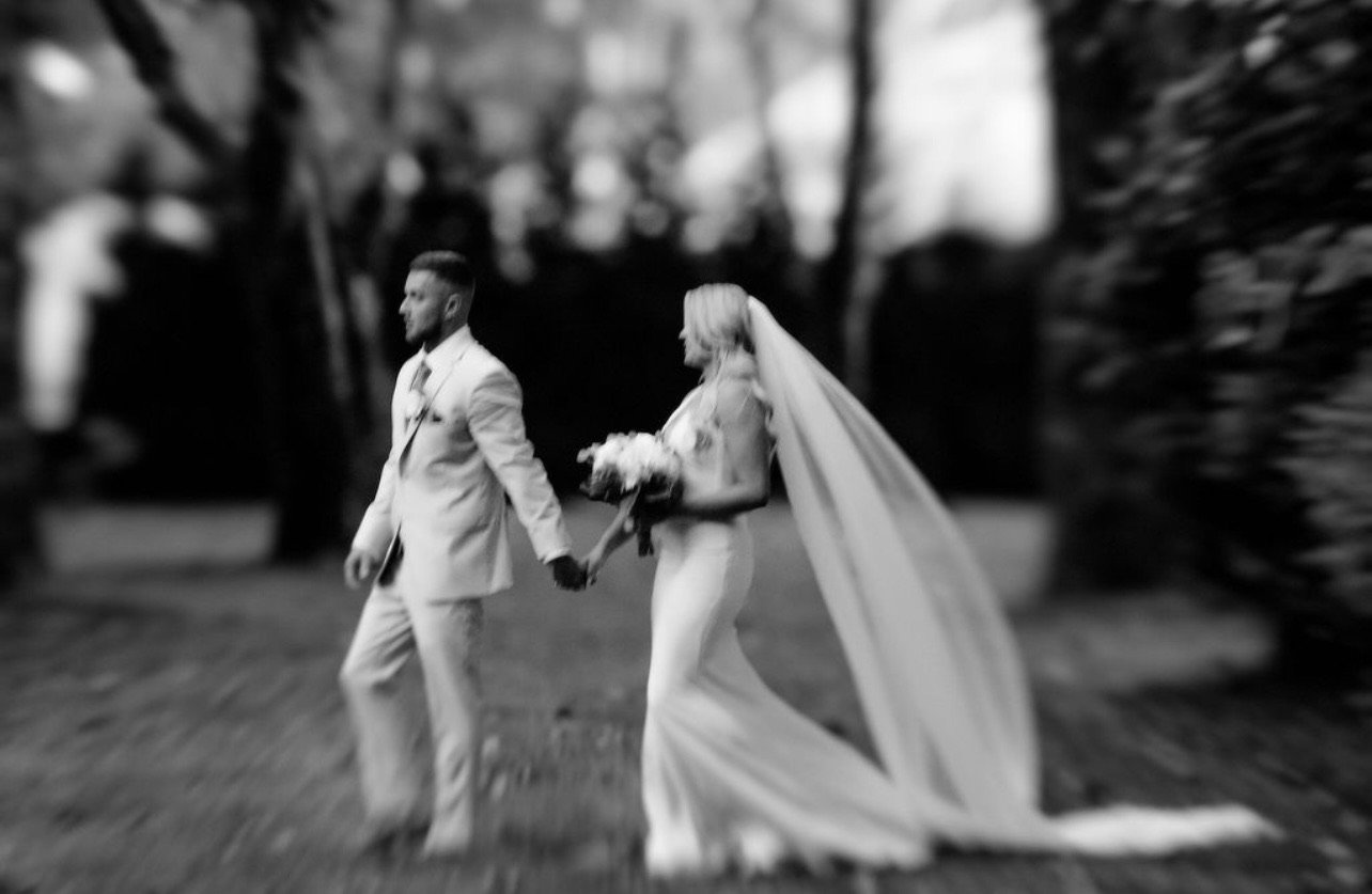 Black and white photo of a bride and groom holding hands and walking outdoors, with the bride carrying a bouquet and wearing a long veil.