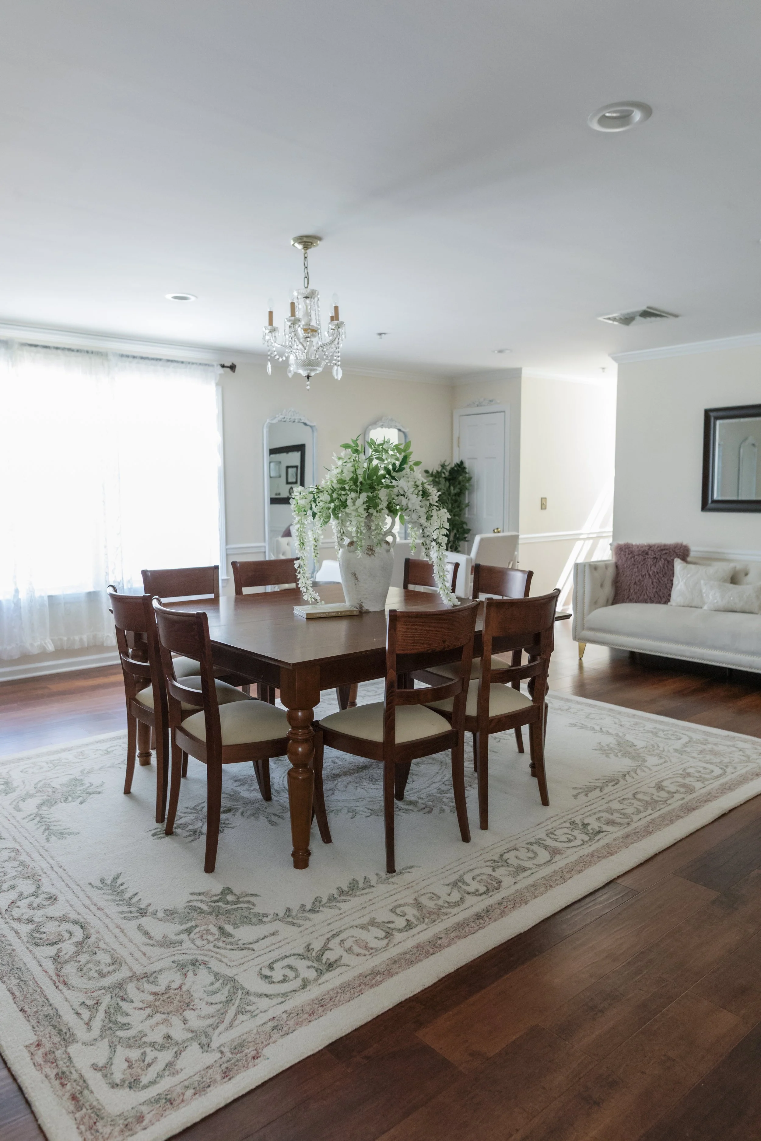A dining room with a wooden table and eight matching chairs, a large floral centerpiece, a chandelier overhead, a window with sheer curtains, a white sofa with cushions, and a decorative mirror on the wall.