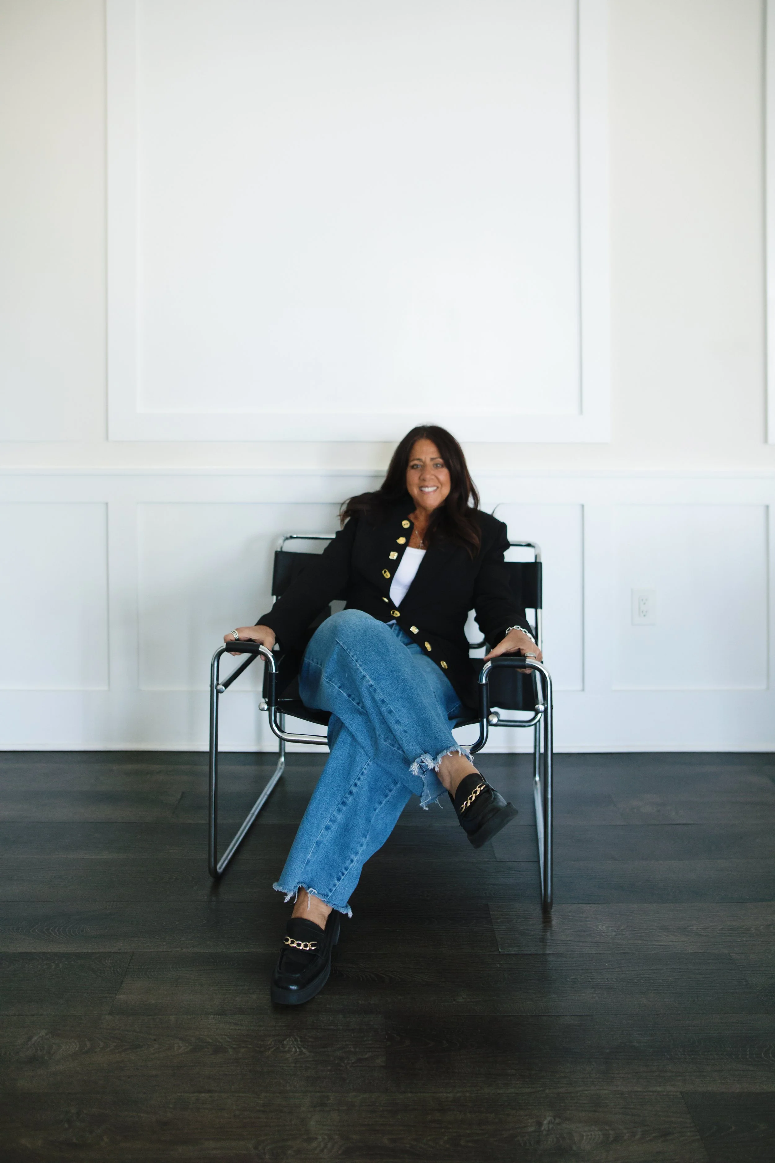Woman sitting on a black chair with arms crossed in a room with white walls and dark wooden floor, smiling at the camera.