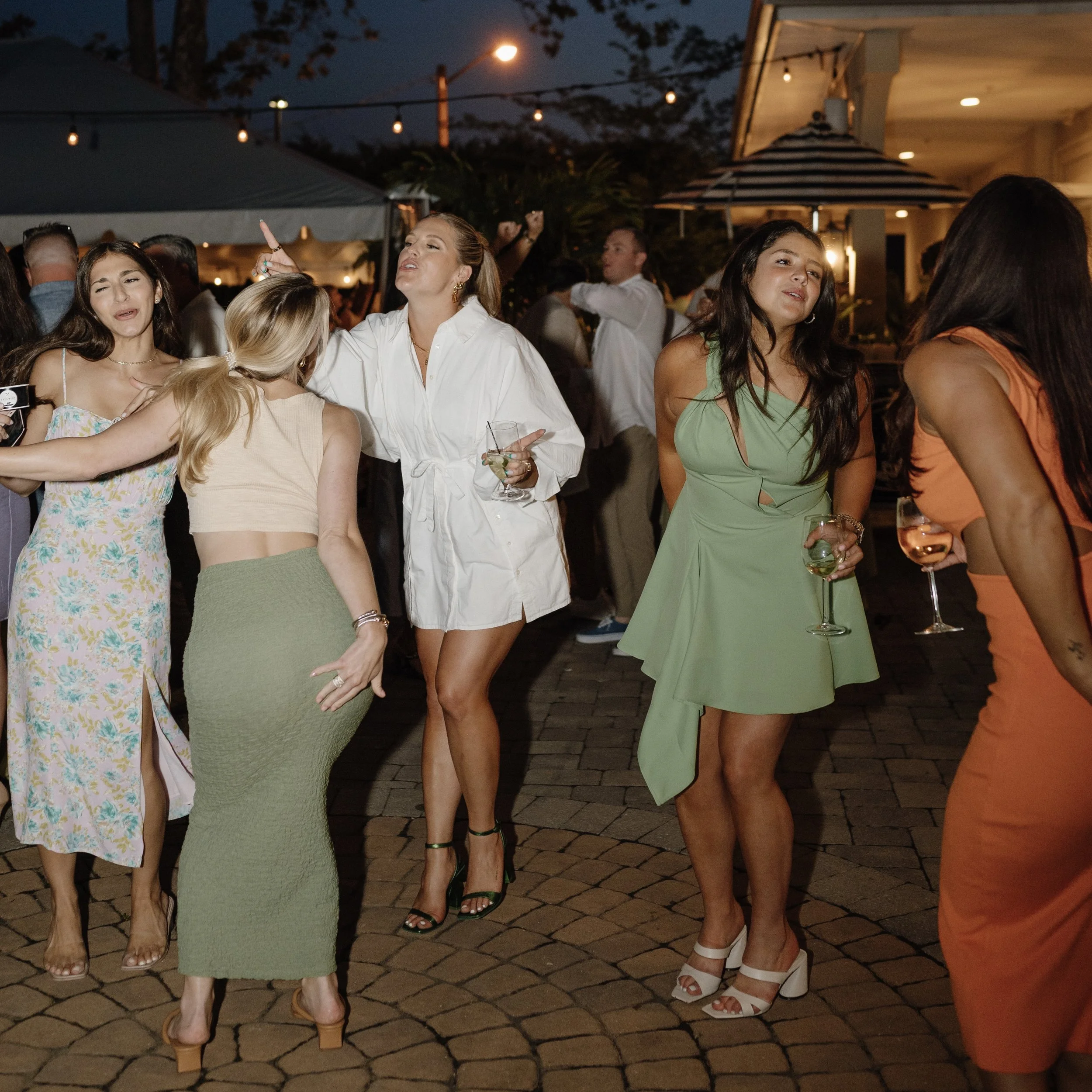 Group of women dancing and socializing at an outdoor evening party with string lights, tents, and a house in the background.