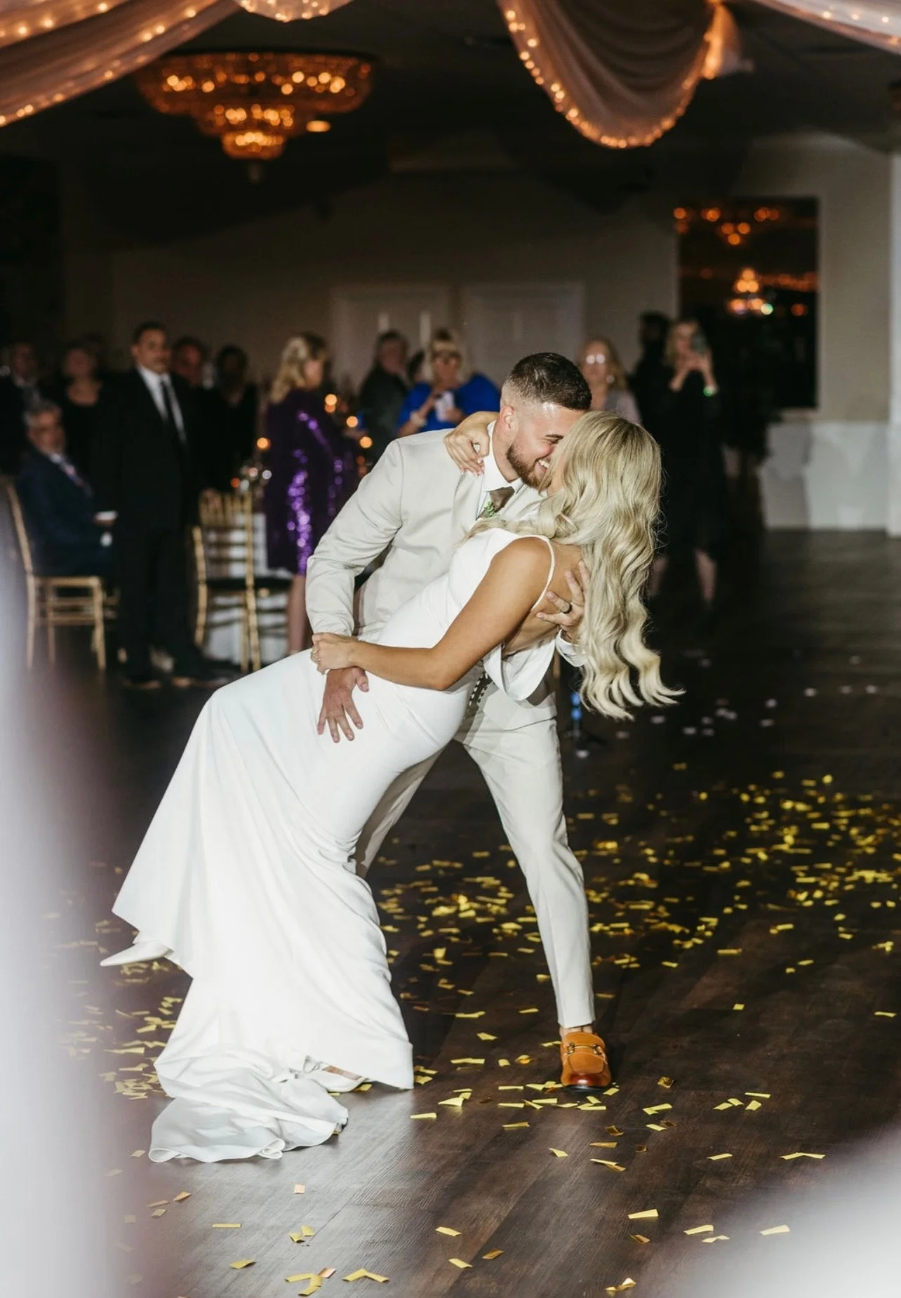 A couple dancing at a wedding reception, with the groom dipping the bride while she leans back. Guests watch in the background, with decorated ceiling and scattered gold confetti on the floor.