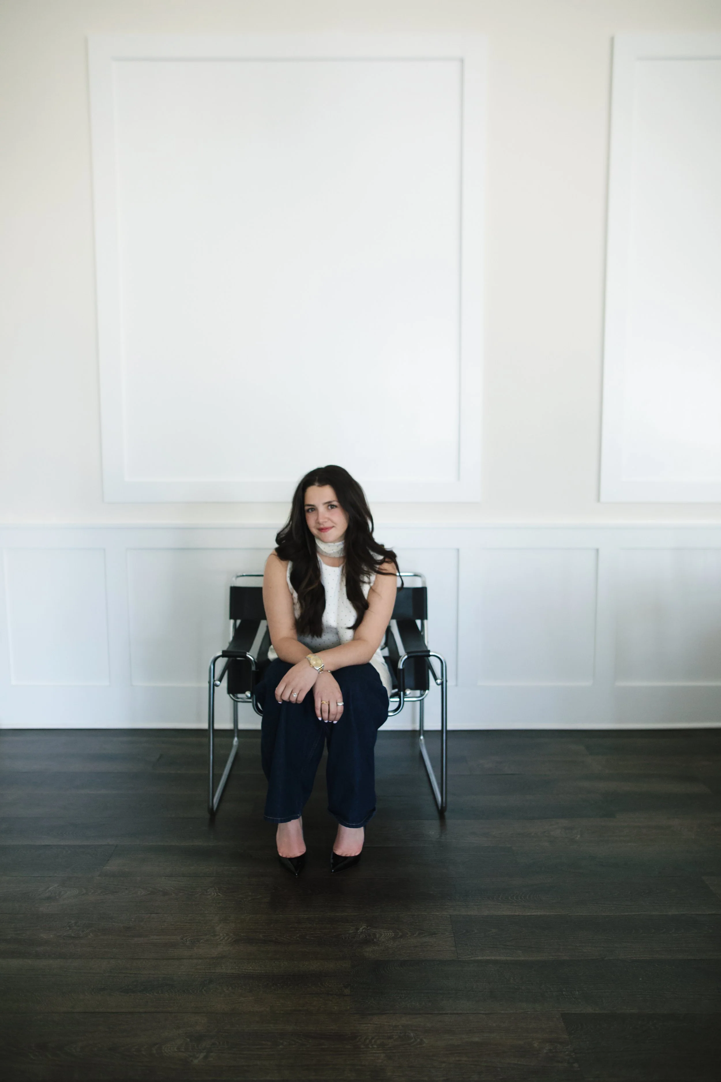 A woman sitting on a black chair in a room with white walls and dark wooden floor.