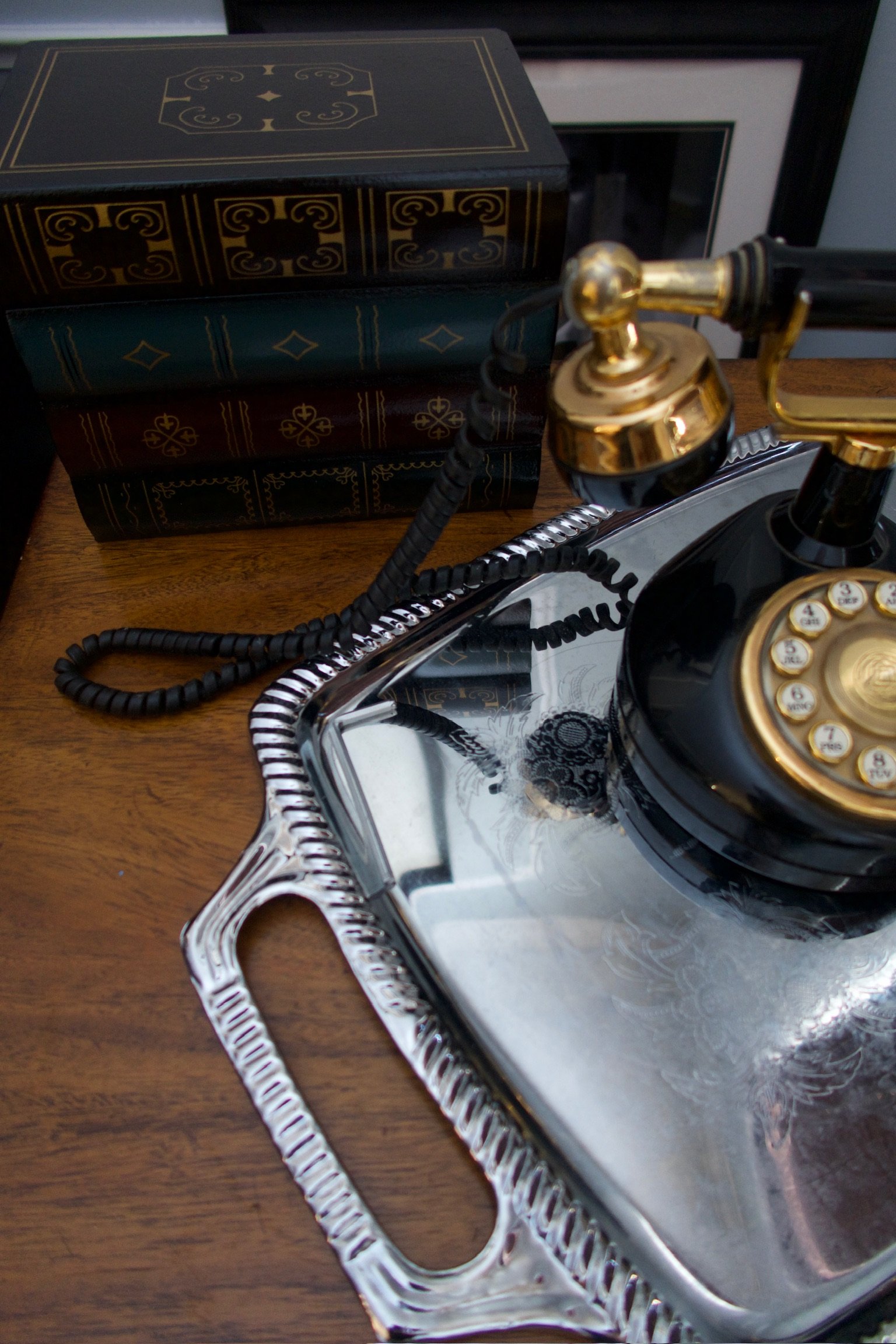 Close-up of a vintage black rotary phone with gold accents on a silver tray, beside a stack of decorated books on a wooden surface.