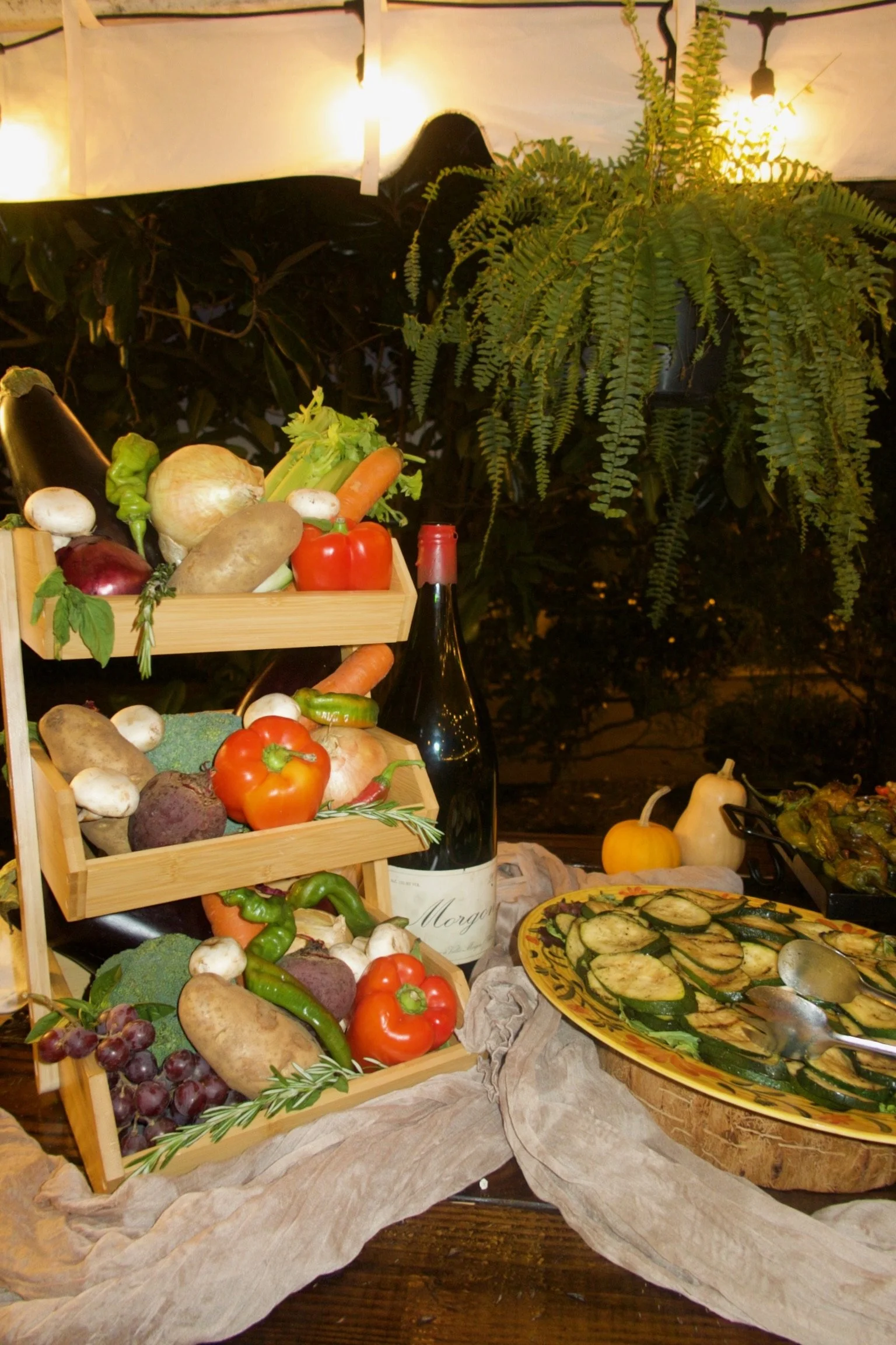 Display of fresh vegetables, a wine bottle, and roasted zucchini slices at a dinner table, with a large fern plant in the background.