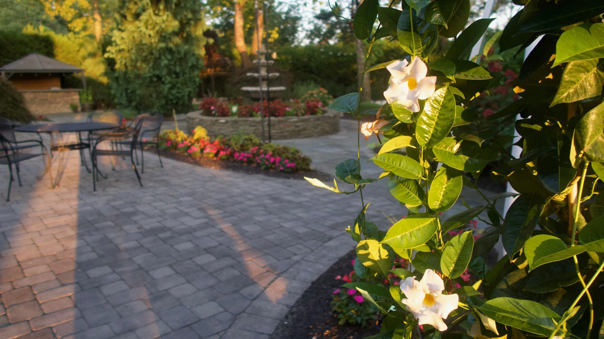 A garden patio with a brick floor, outdoor black metal tables and chairs, flower beds with pink and yellow flowers, a raised flower bed with red flowers, and a small gazebo in the background, all illuminated by warm sunlight, with greenery and trees 