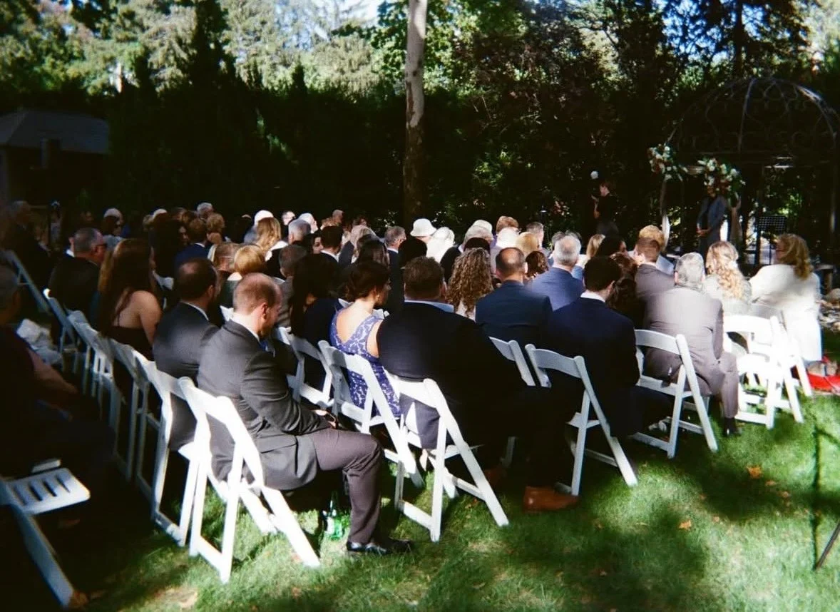 People attending an outdoor wedding ceremony, seated on white chairs on a grassy area, with a wedding arch decorated with flowers in the background. Tall trees and dense foliage surround the scene.