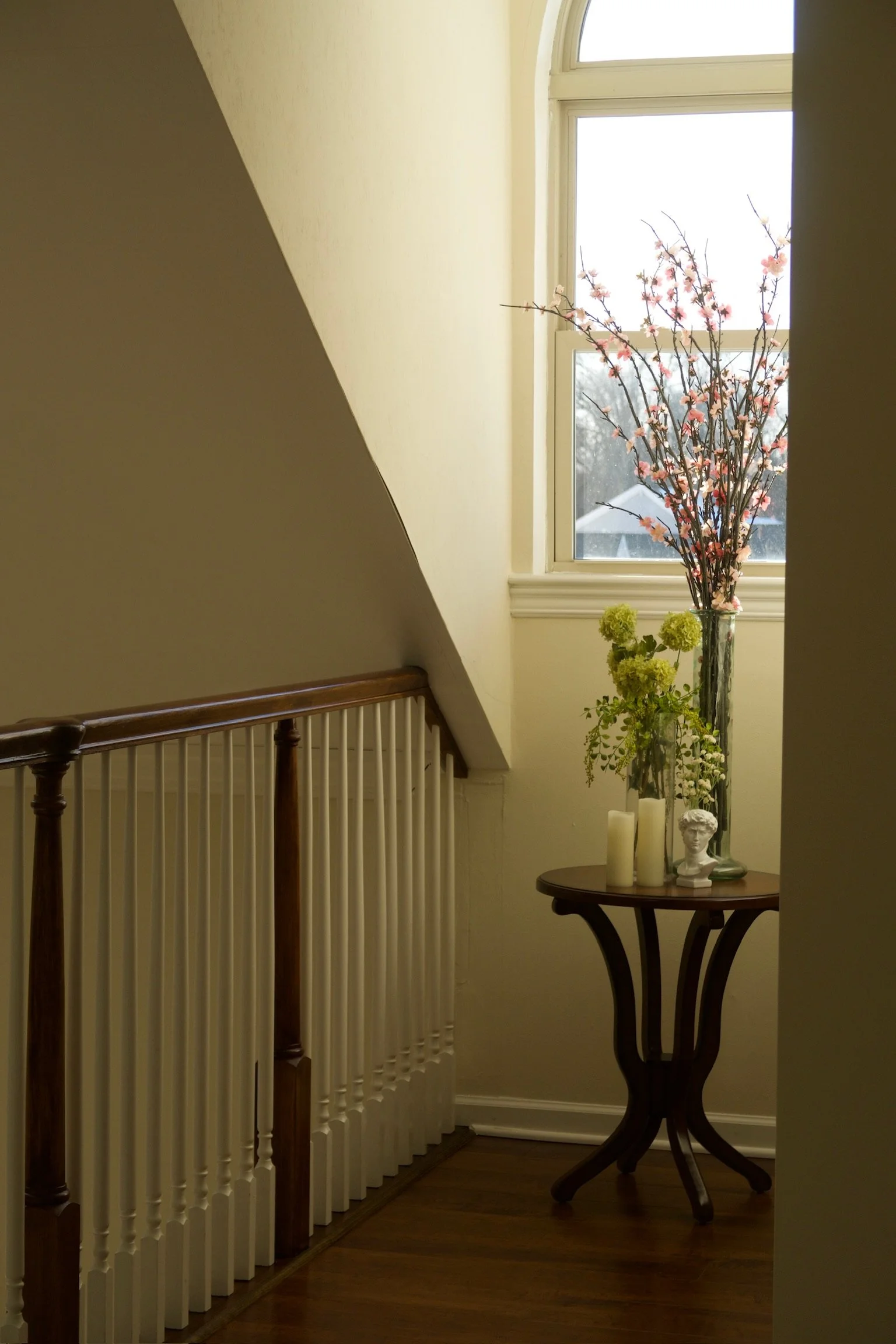 Decorative table with candles, a small bust, and flower arrangements near a window with pink blossoms outside