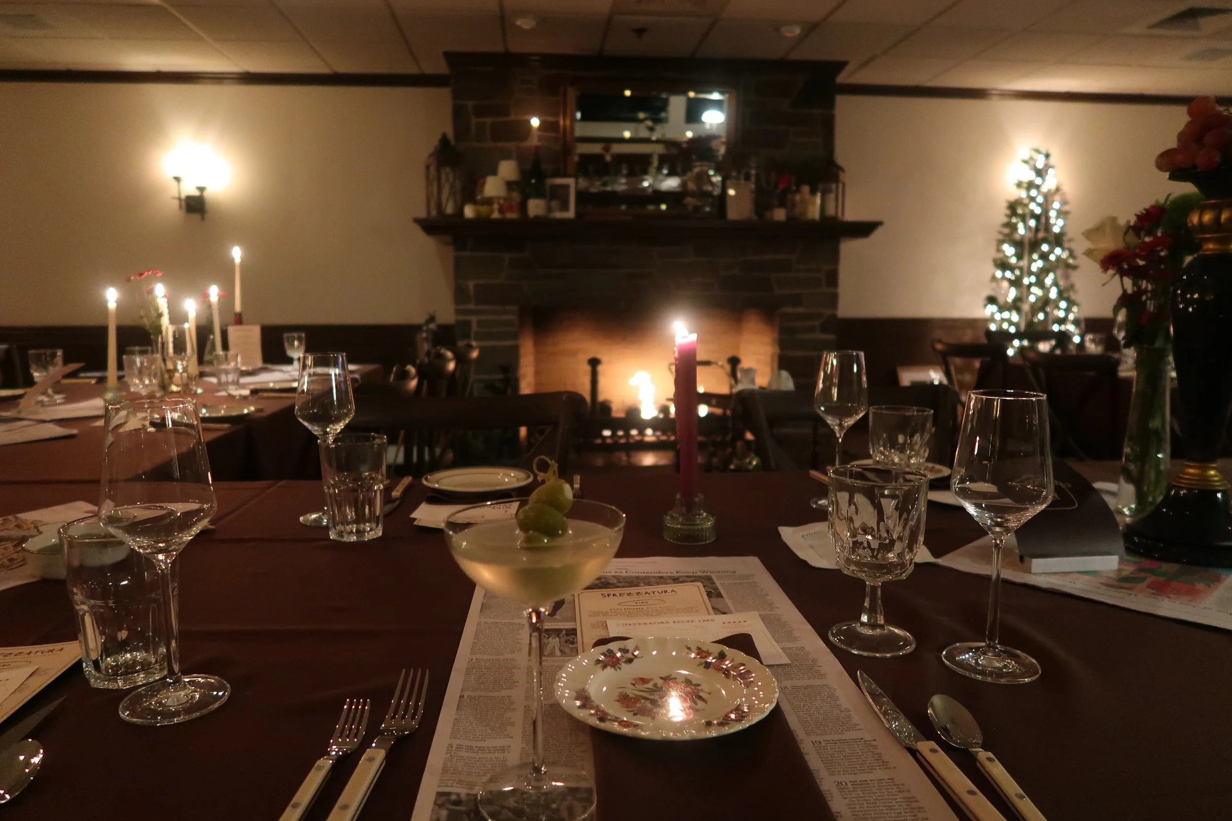 A cozy dining room with a long table set for a meal, lit by candlelight and a fireplace, decorated for the holiday season with a small Christmas tree and flowers.