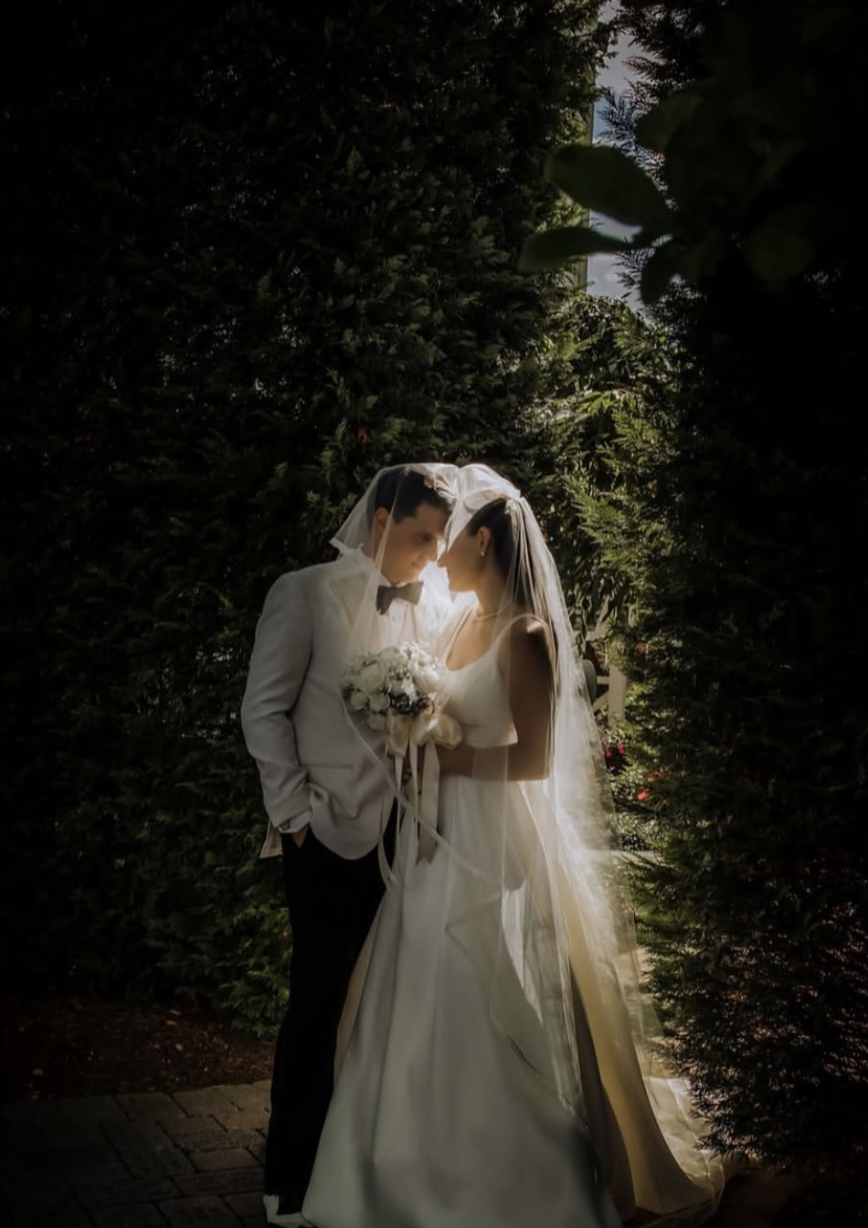 A bride and groom are standing close together outdoors, under a veil, with lush greenery surrounding them. The bride is holding a bouquet of white flowers, and they are about to touch foreheads in a romantic pose, with sunlight filtering through the 