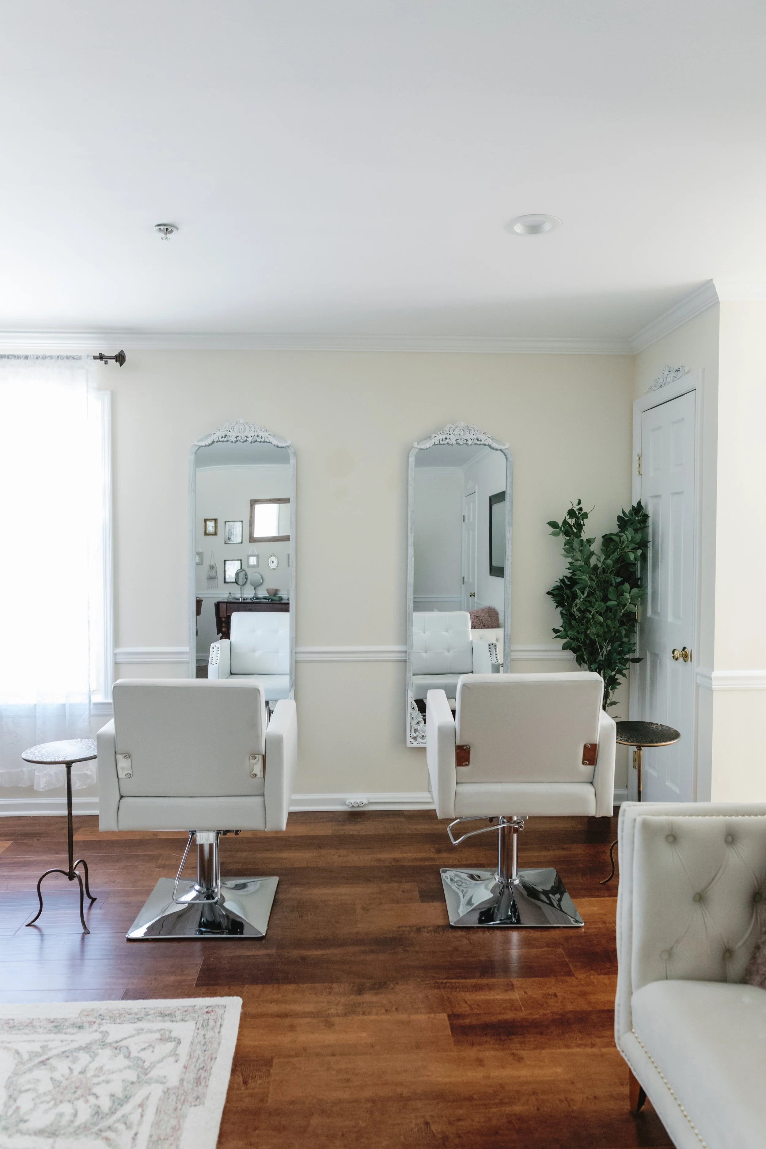 A room in a beauty salon or barber shop with two white styling chairs in front of two tall mirrors and a small side table on the left. There is a person sitting in the reflection of the mirrors. The room has cream-colored walls, a wooden floor, and a large potted plant near a white door.