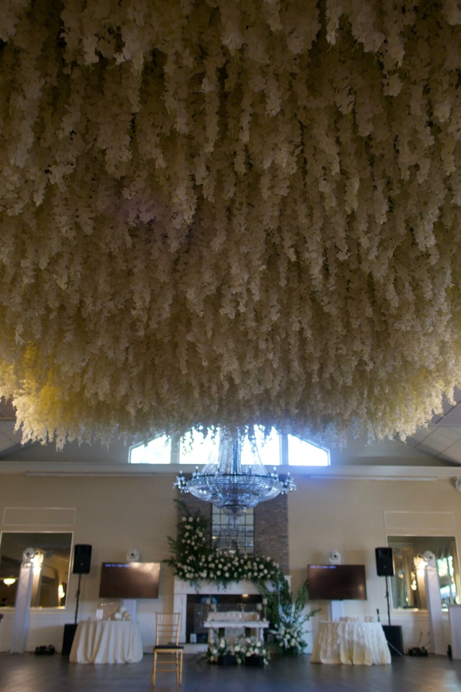 Interior of a decorated venue with a large floral ceiling installation and a chandelier, floral arrangements, and tables below, possibly for a wedding or event.