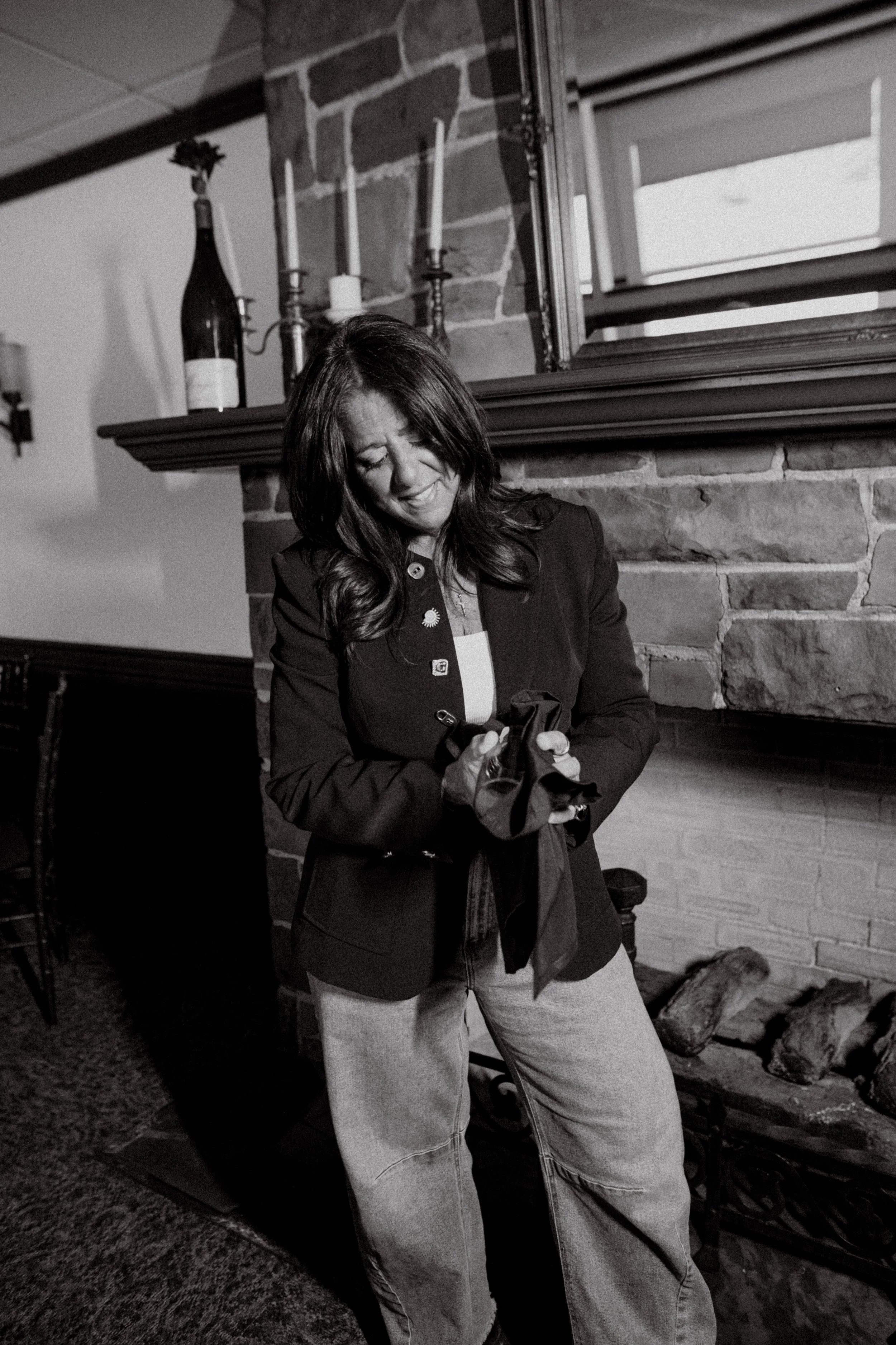A woman with long dark hair, wearing a dark blazer and light jeans, standing indoors near a brick fireplace and smiling while looking into a small bag.