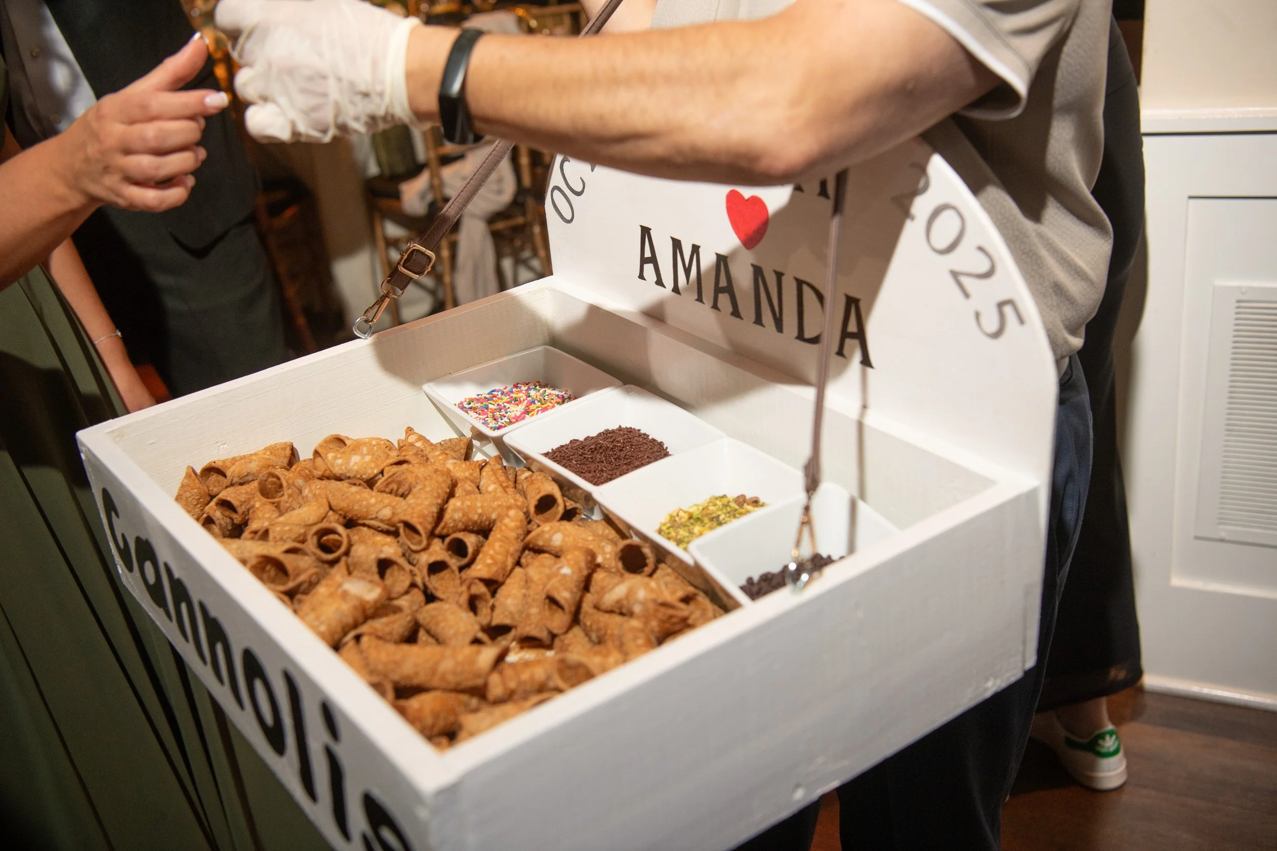 A person handing a box of assorted party favors, including mini cannoli cookies, sprinkles, and chocolate shavings, with a sign saying "Amanda".
