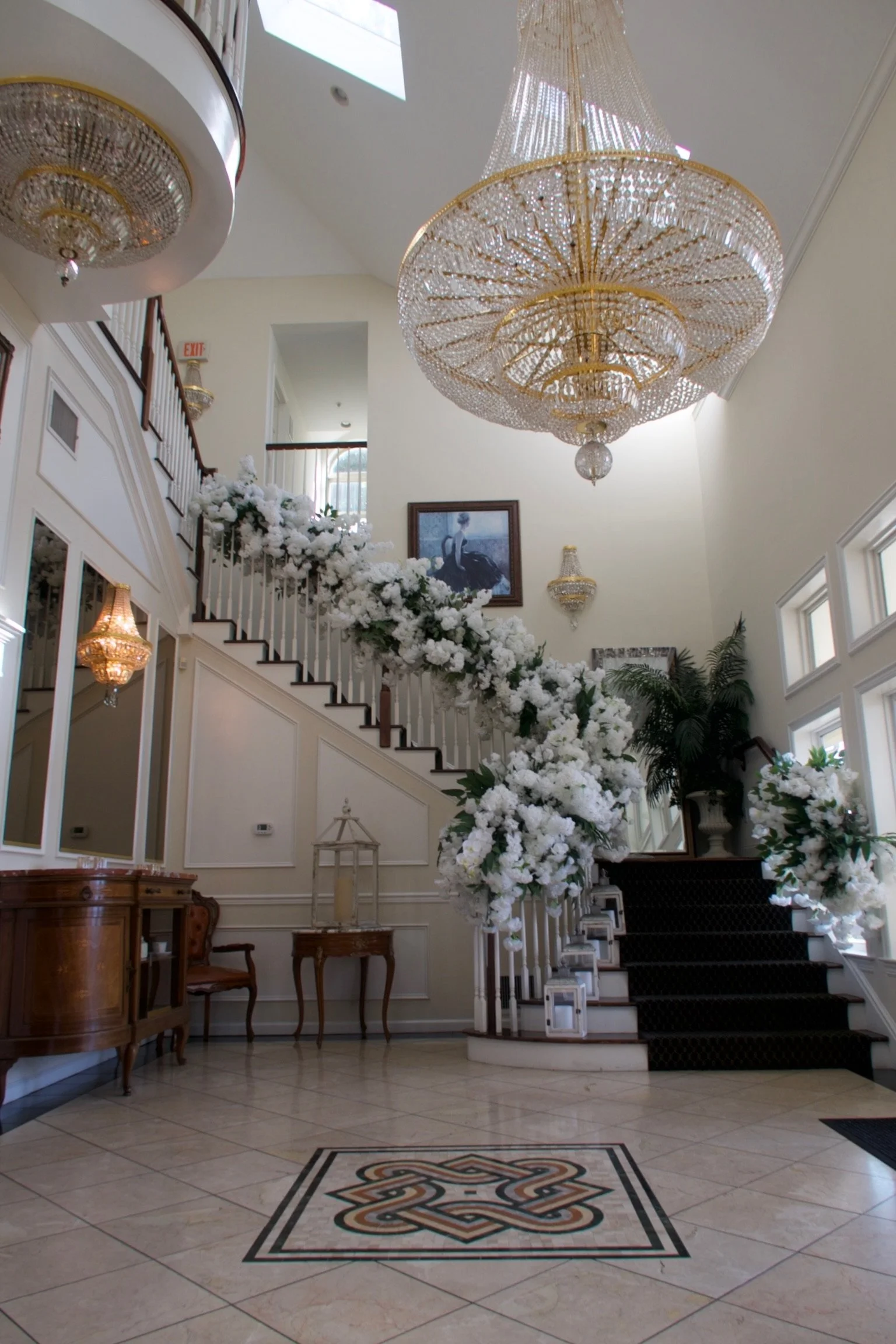 Elegant hotel lobby with a grand staircase decorated with white flowers, large chandeliers, and framed art on the wall.