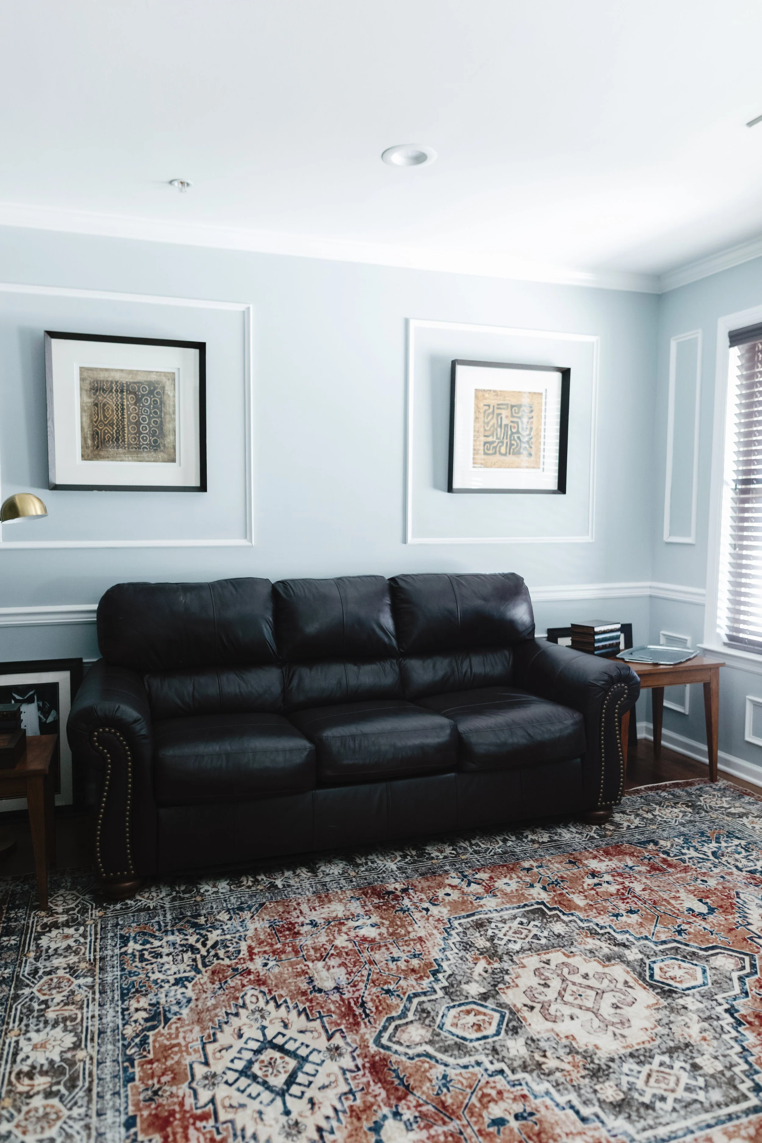Living room with light blue walls, black leather sofa, framed abstract artwork on walls, wooden side tables, patterned area rug, window with blinds.