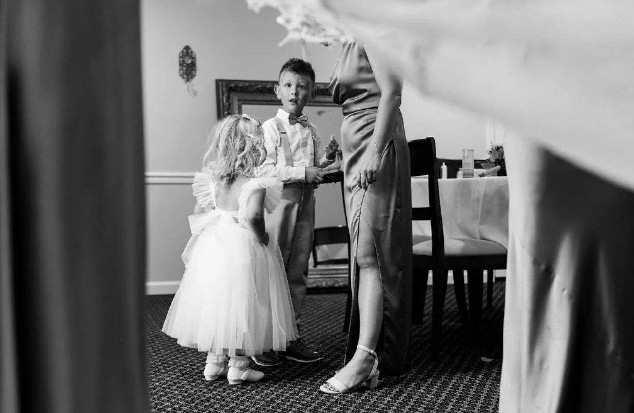 A woman and two children, a girl and a boy, are indoors, dressed in formal attire, with the woman in a satin gown, the girl in a fluffy dress, and the boy in a shirt, suspenders, and bow tie, likely preparing for a special event or celebration.