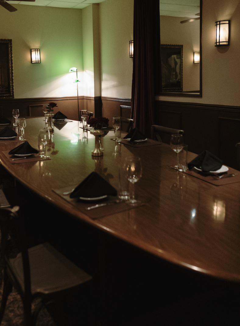 A formal dinner table set with black napkins, glassware, and floral centerpieces in a dimly lit room with wall sconces and a large mirror.