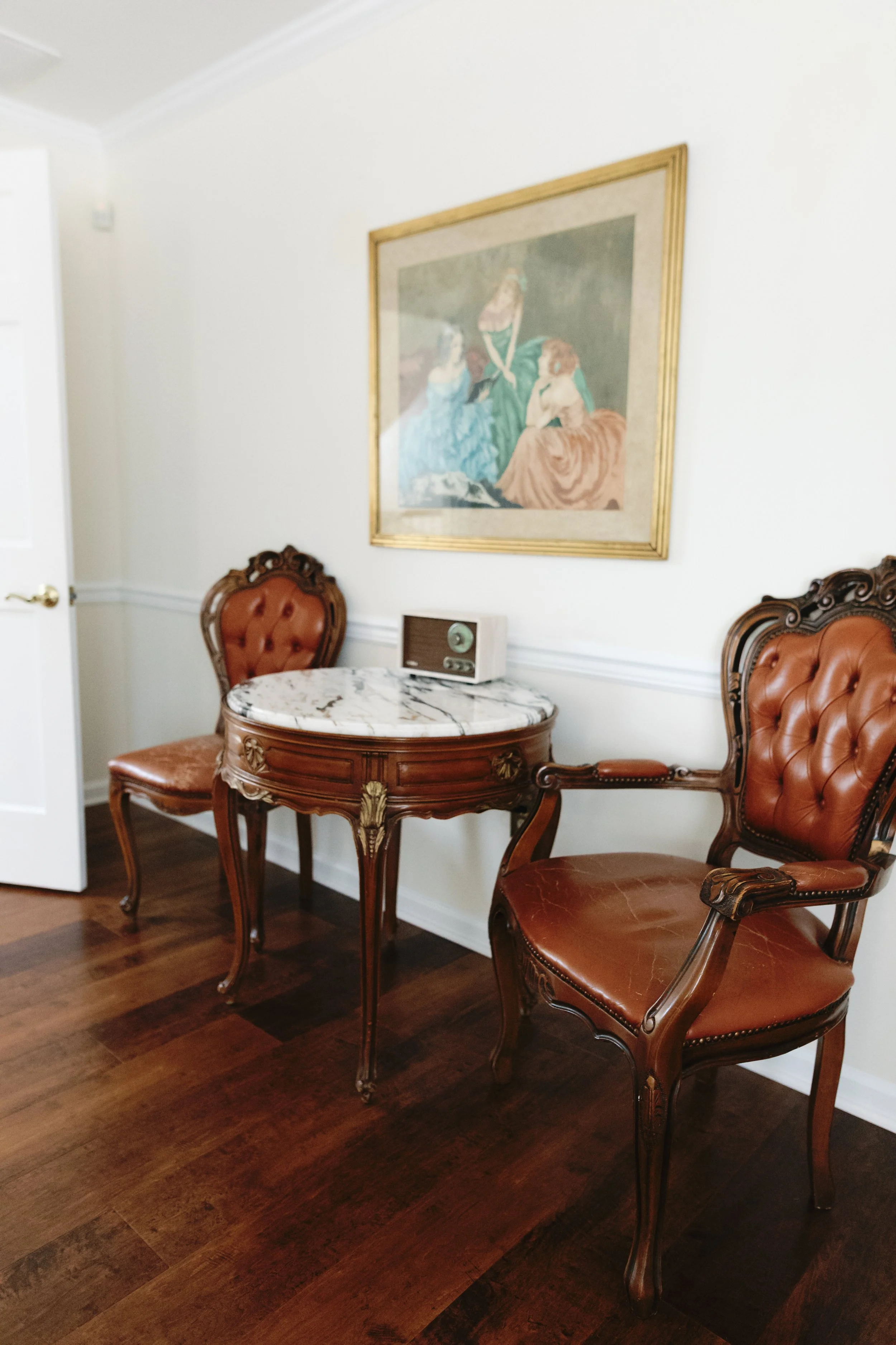 Vintage interior with two ornate wooden chairs with leather upholstery, a marble-topped side table, a framed painting on white walls, and hardwood flooring.