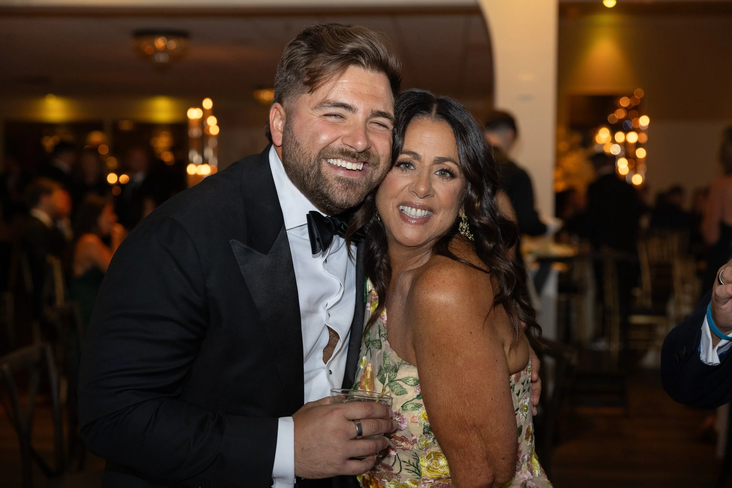 A man in a tuxedo and a woman in a floral dress smiling and hugging at a formal event.