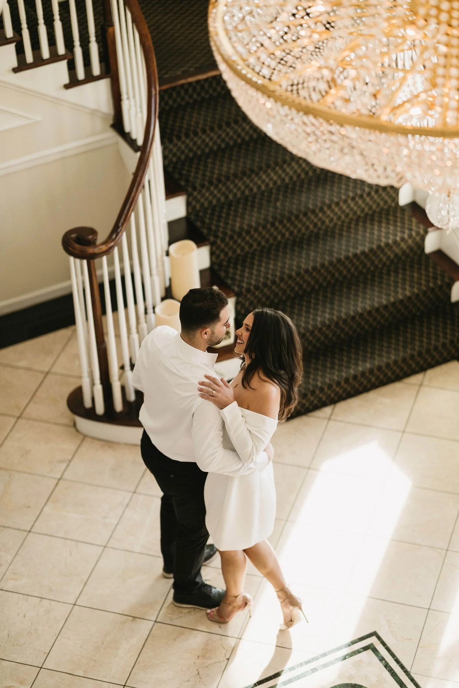 A couple dancing indoors under a chandelier, with a staircase nearby.