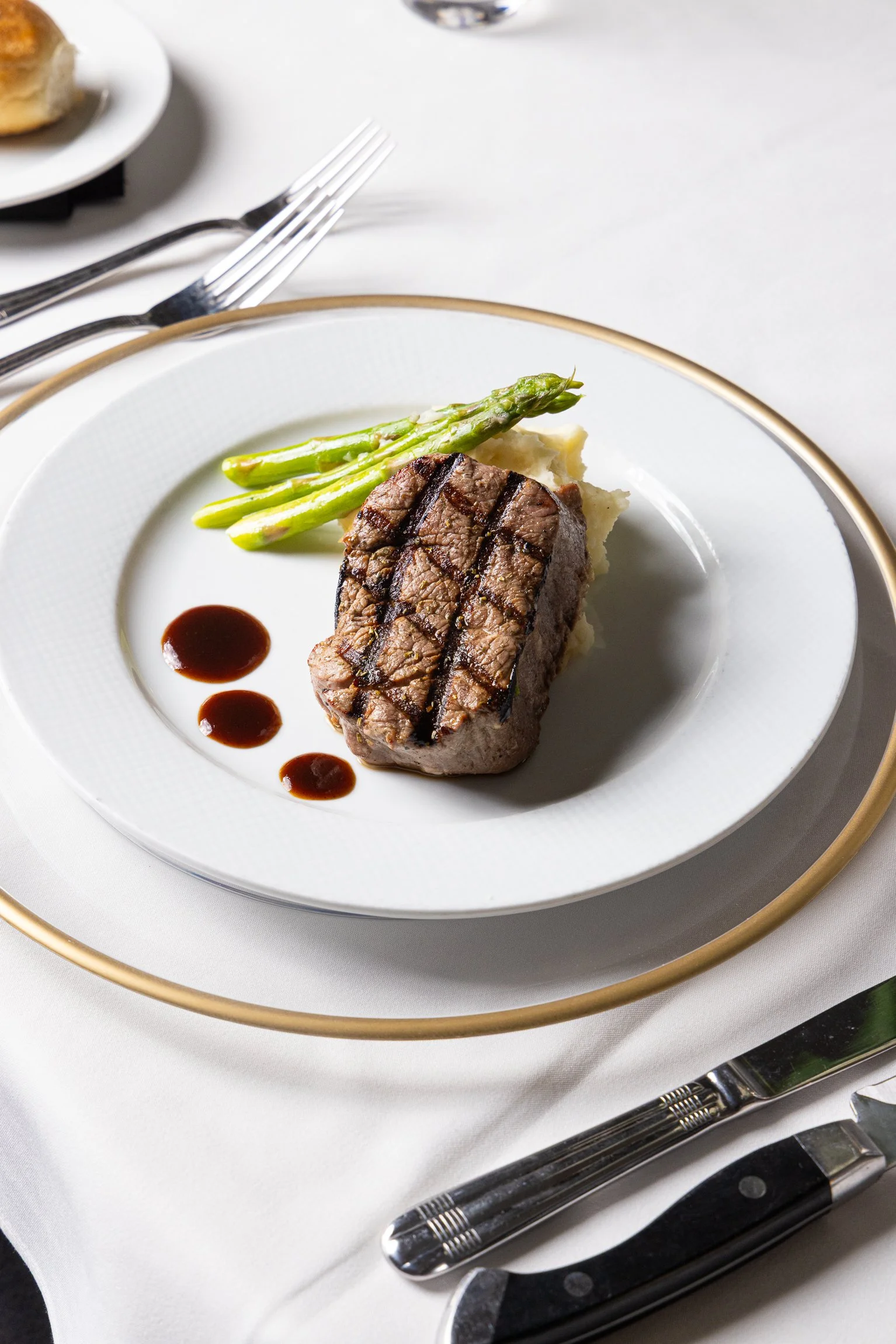 A plate with grilled steak, asparagus, mashed potatoes, and sauce dots on a white tablecloth.