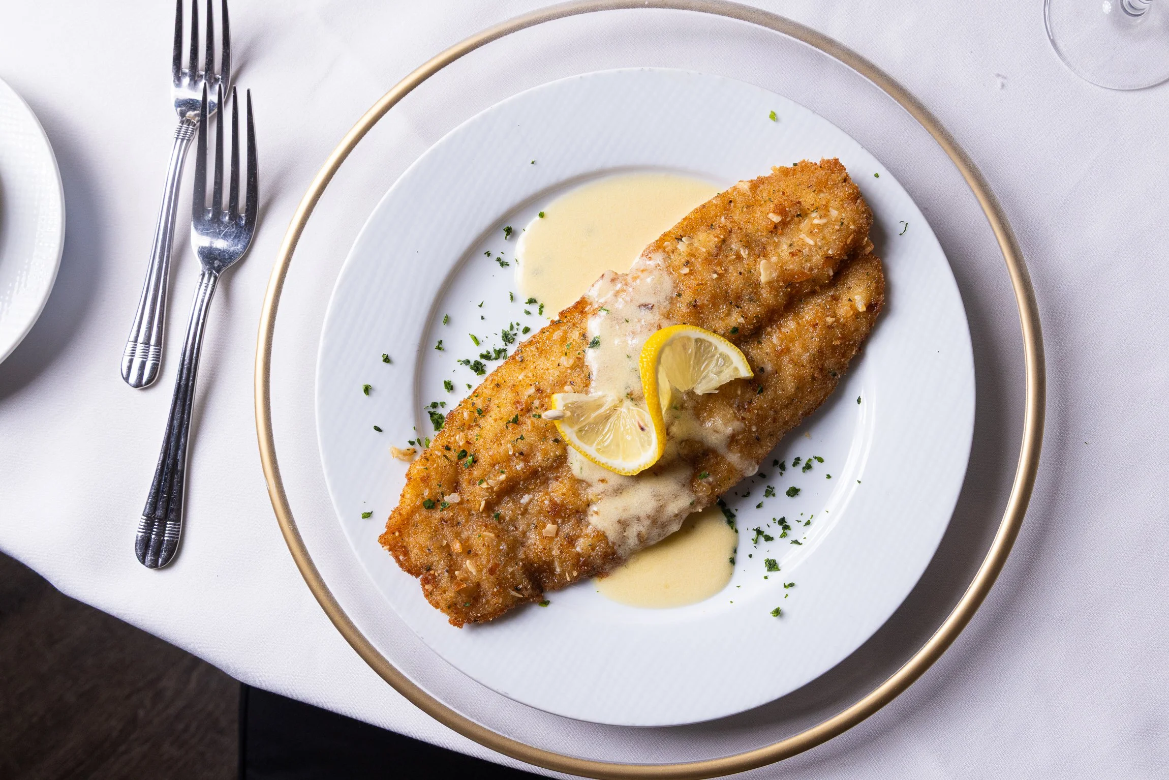 Breaded and fried fish fillet topped with lemon slices and cream sauce, served on a white plate with parsley garnish, on a white tablecloth.
