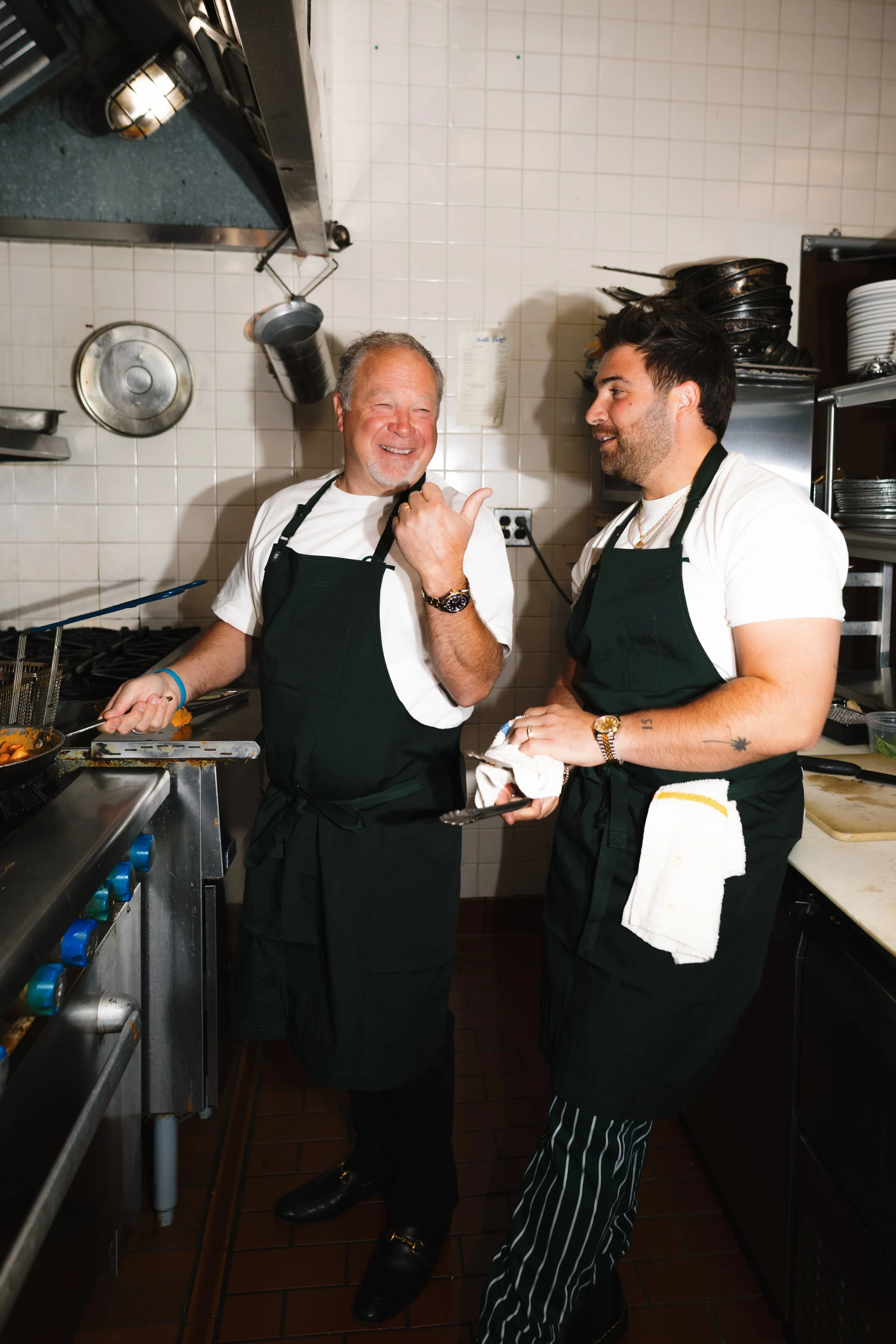 Two male chefs in black aprons smiling and talking in a professional kitchen. One is an older man with gray hair, the other a younger man with dark hair. They are surrounded by kitchen utensils, pots, and plates.