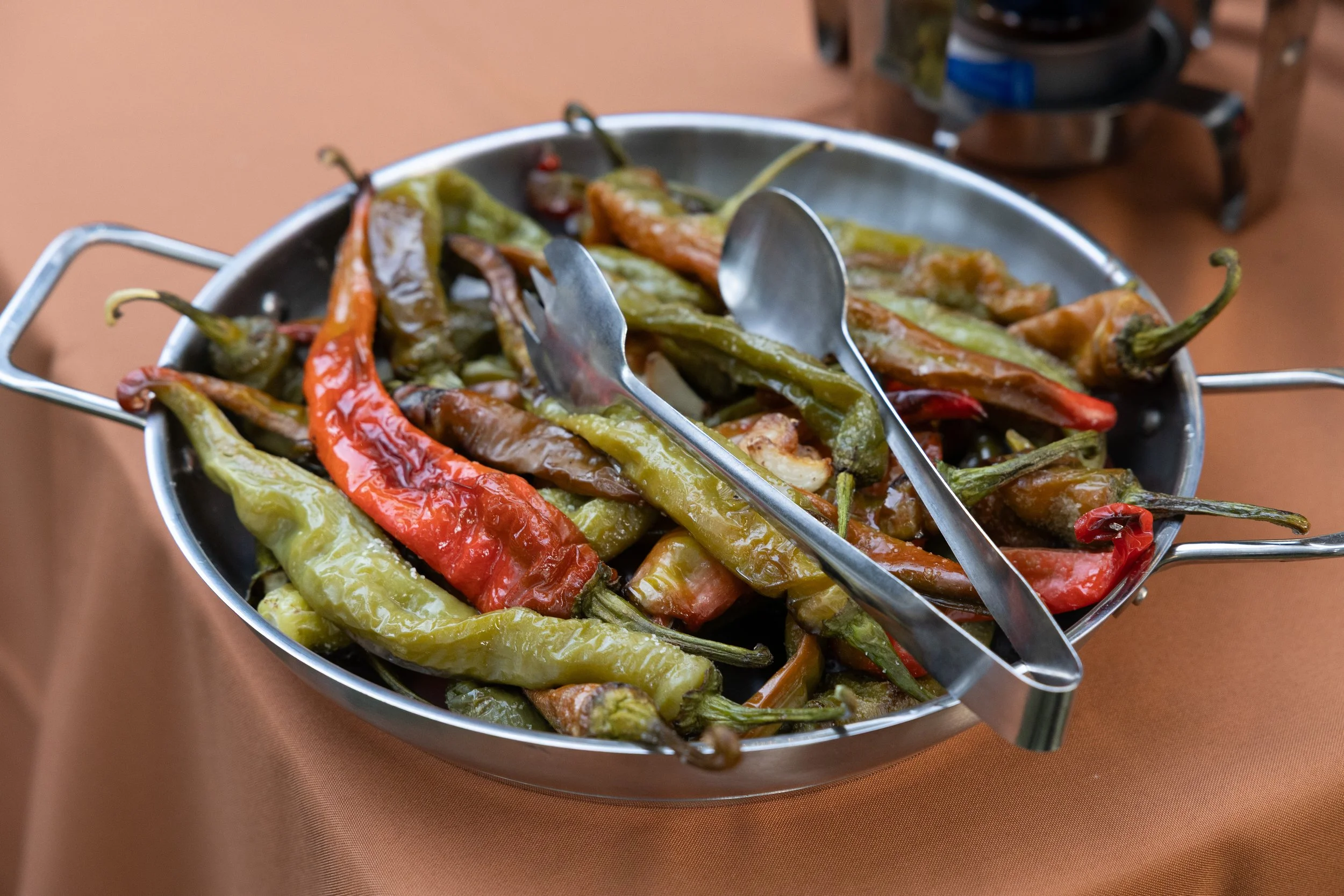 A metal platter filled with roasted chili peppers, some red and some green, with metal tongs and a serving spoon on top, placed on a brown table.