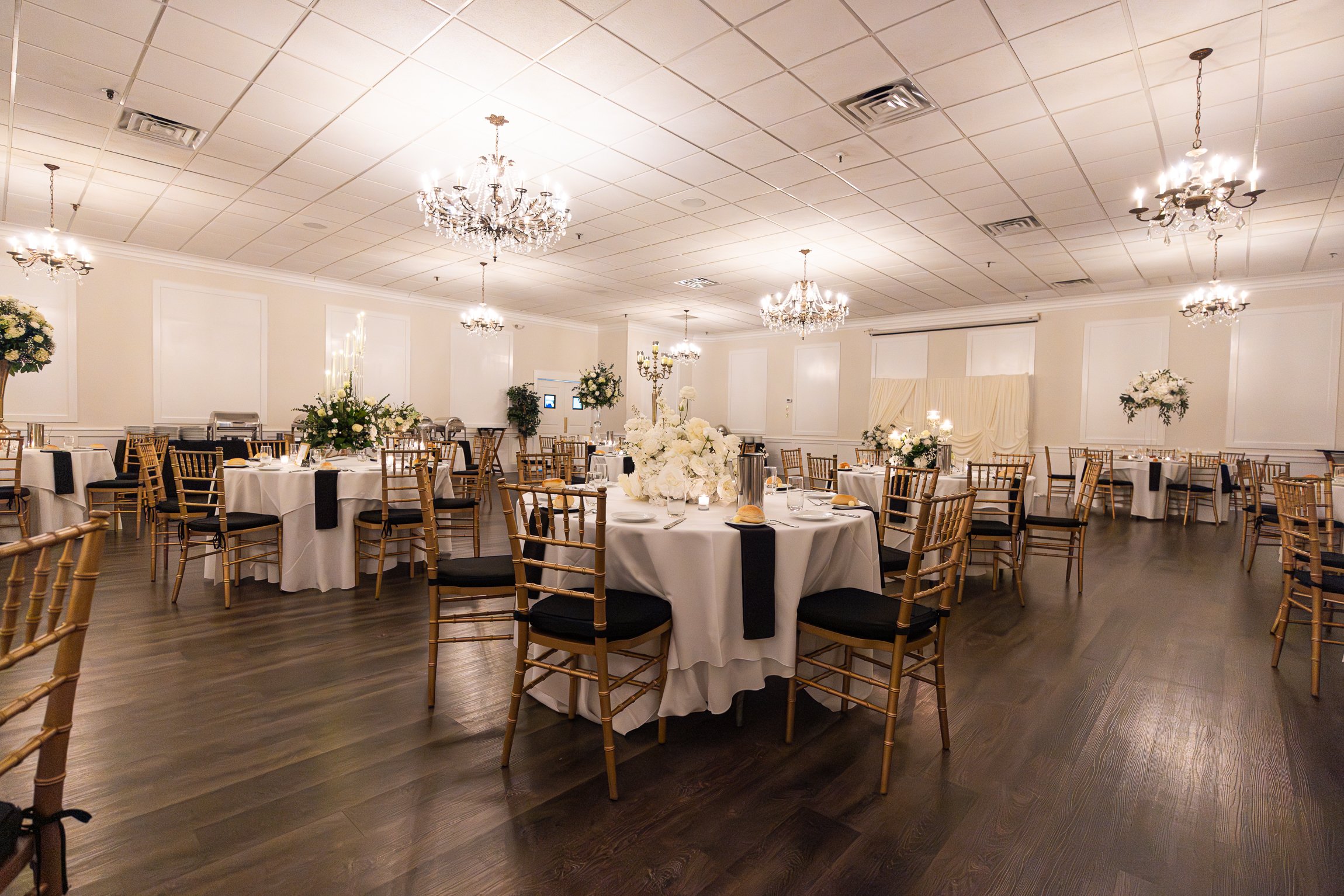 Elegant banquet hall decorated for a wedding or formal event with round tables, gold chairs, white tablecloths, floral centerpieces, chandeliers, and soft lighting.