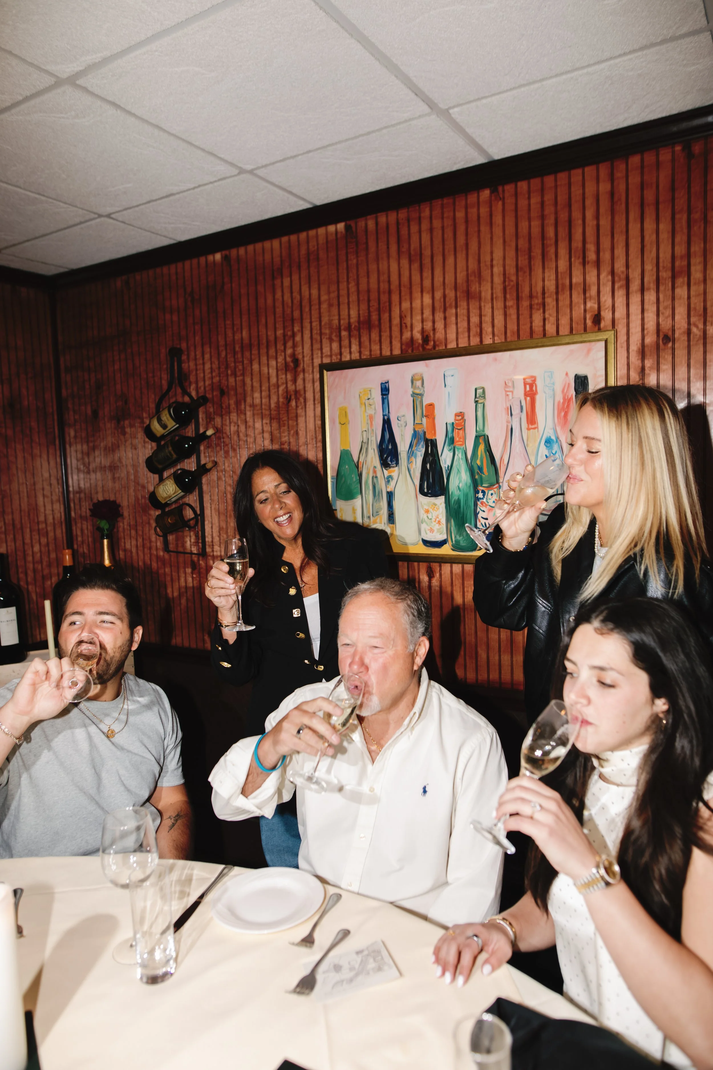 Group of five people celebrating with wine at a restaurant, with artwork of bottles on the wall behind them.
