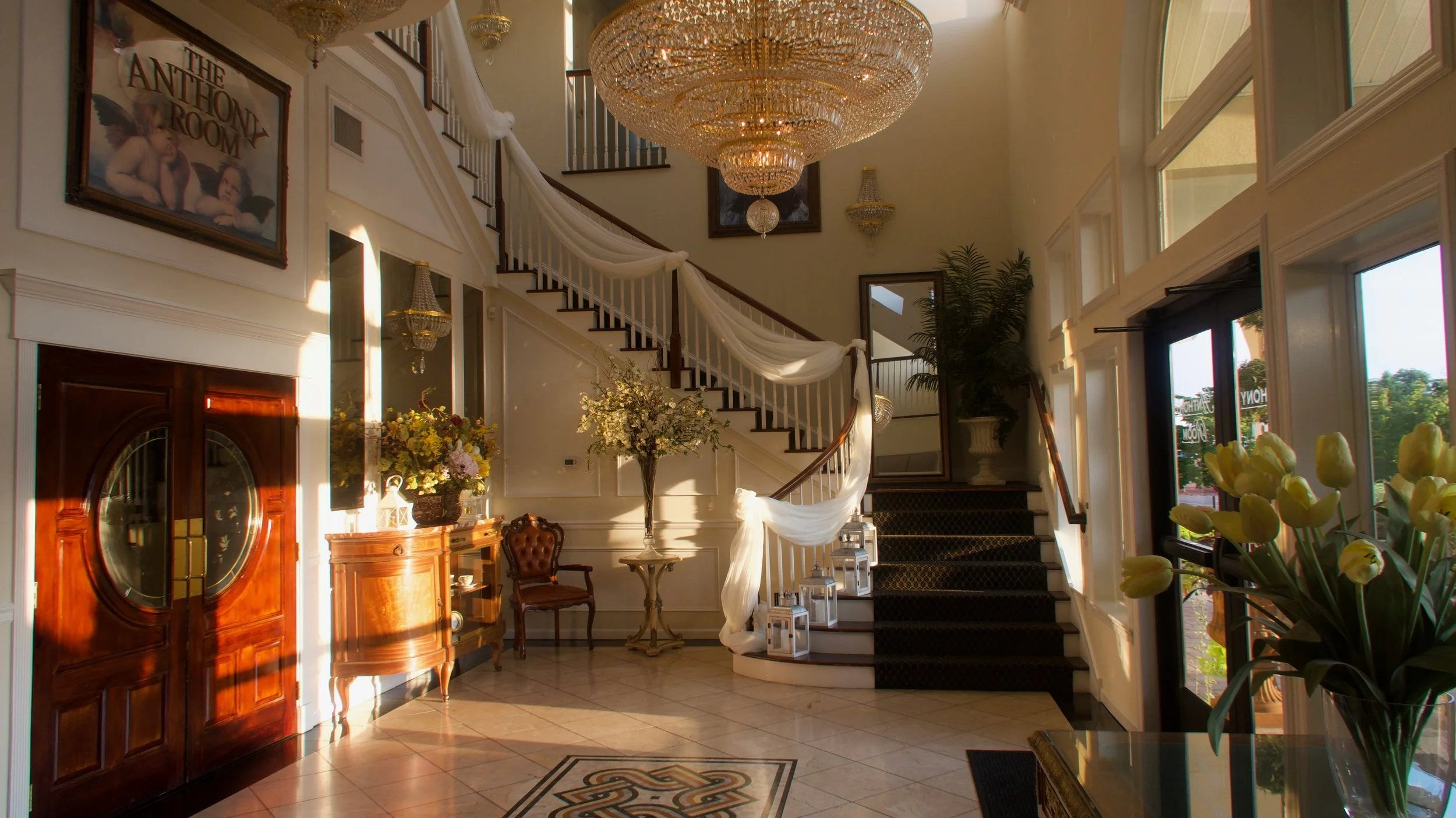 Elegant hotel lobby with grand staircase decorated with white drapery, large chandelier, wooden furniture, and floral arrangements.