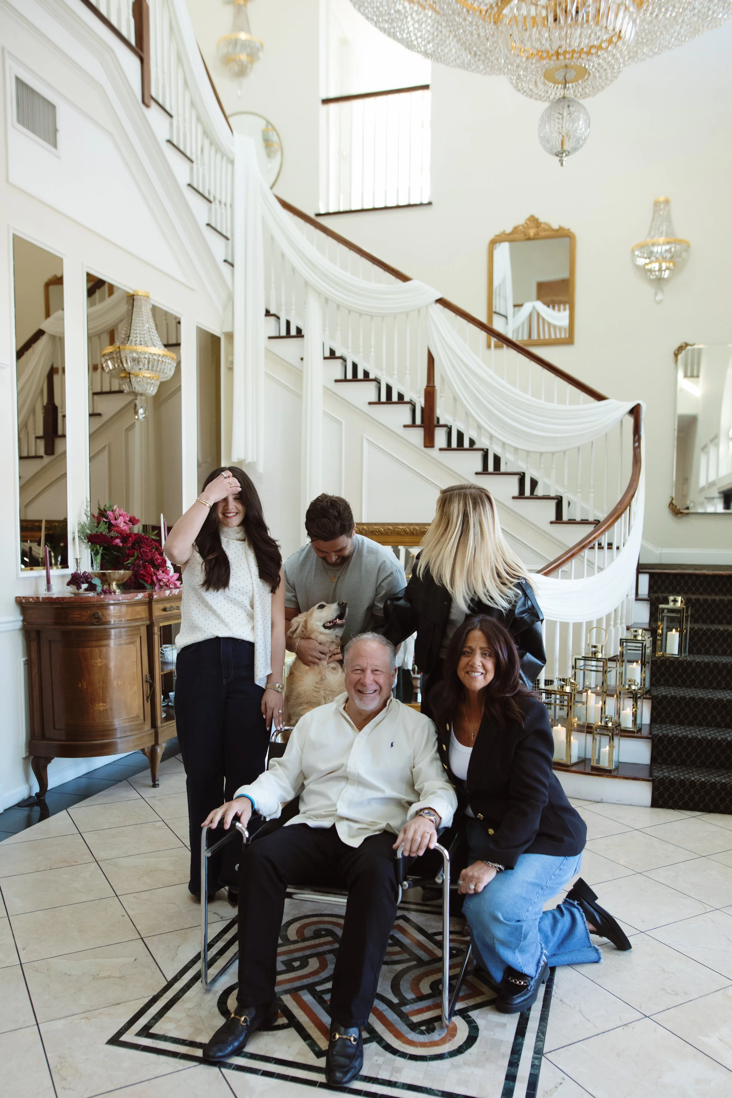 A group of six people and a dog gathered in a luxurious entryway with a staircase, chandelier, and decorative lanterns.