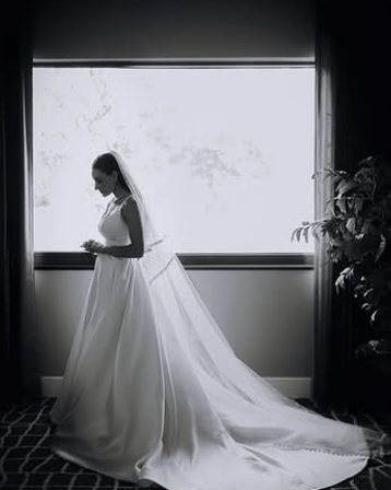 Bridal gown on a bride standing by a large window with curtains and plants.
