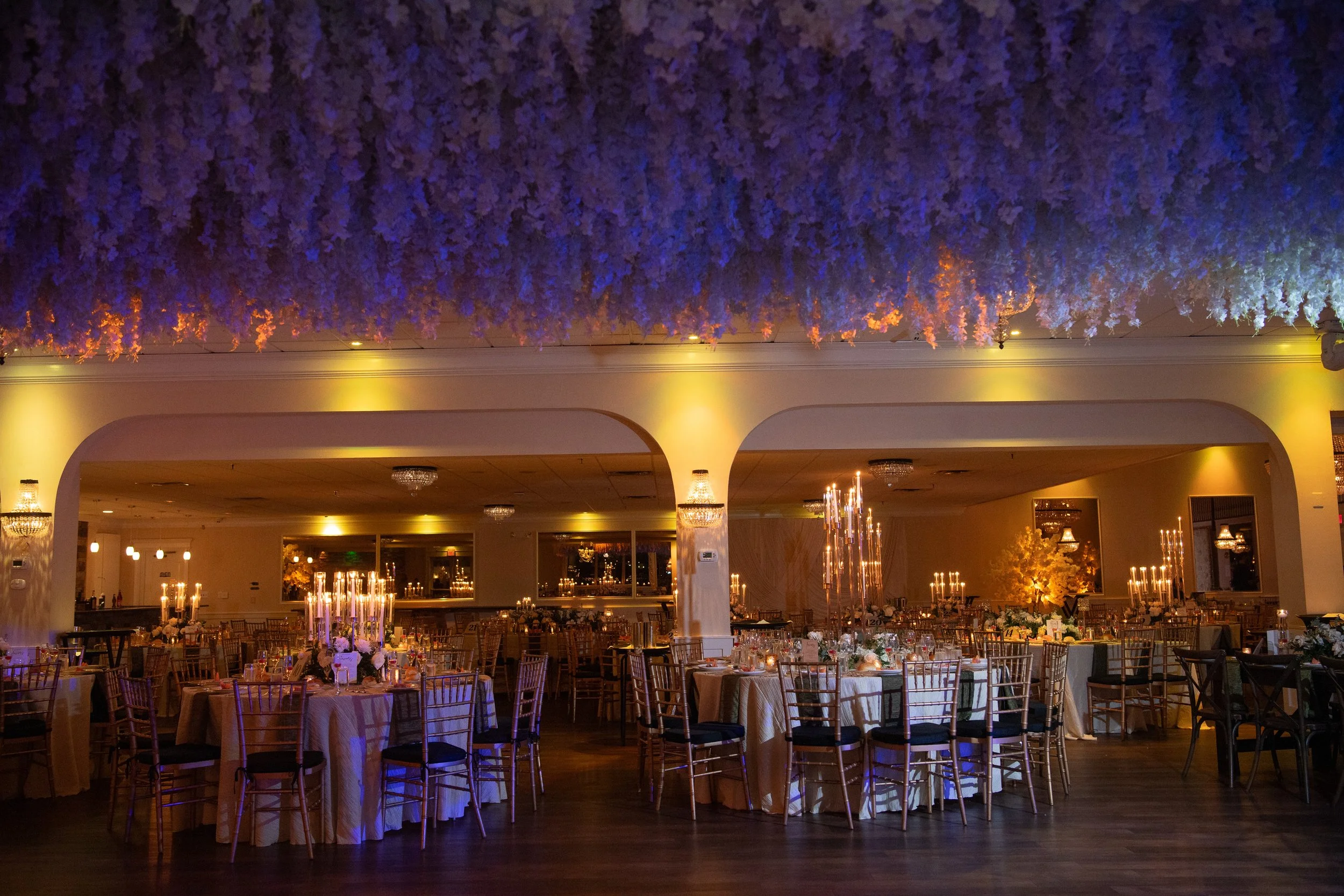 Elegant indoor banquet hall decorated for a wedding reception with round tables, gold chairs, white tablecloths, floral centerpieces, and multiple candleholders with lit candles; ceiling adorned with hanging purple flowers and soft warm lighting.