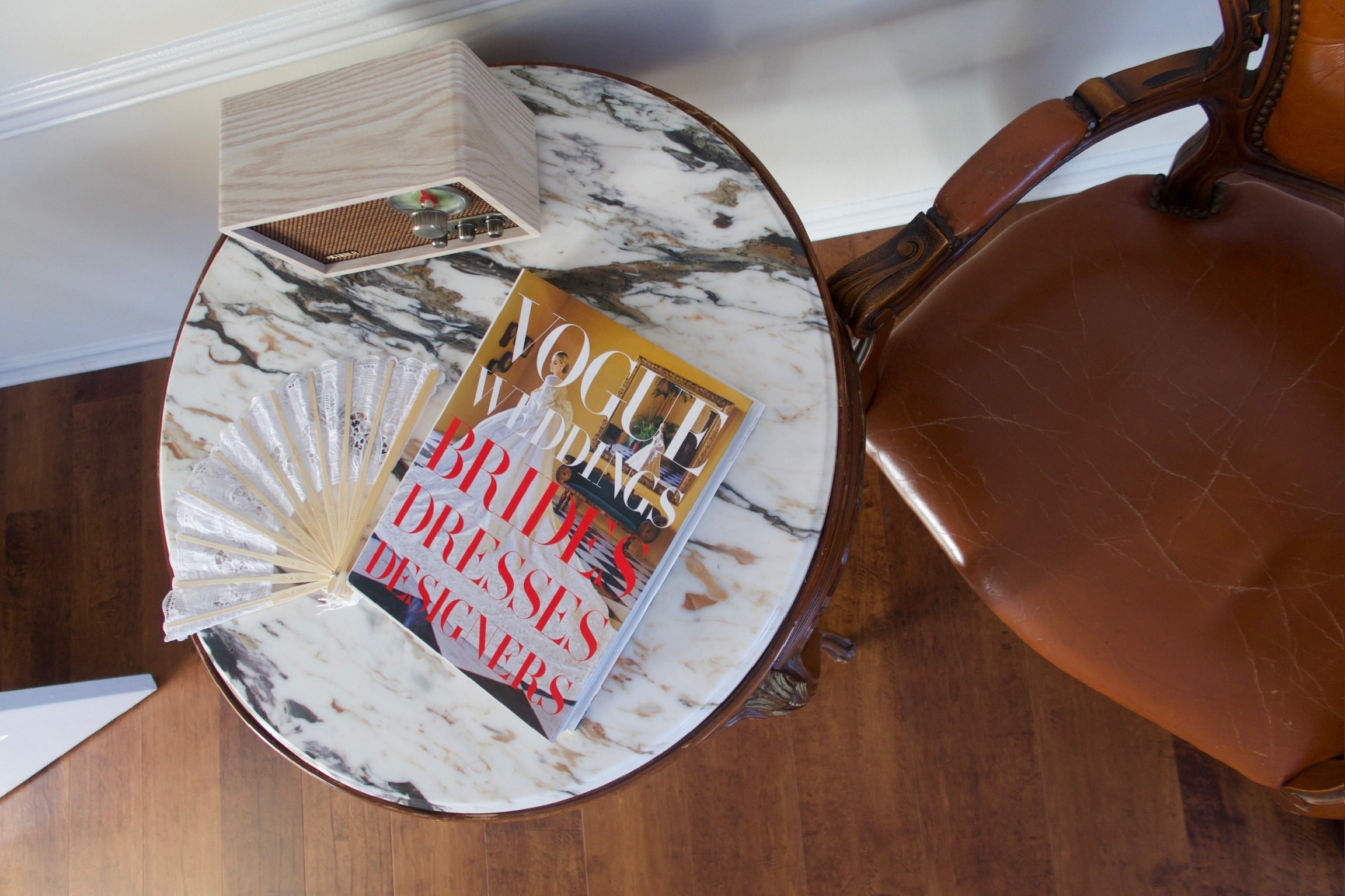 Top-down view of a marble-top table with a Vogue magazine, a fan-shaped decorative object, and a box on it, next to a vintage brown leather armchair on a wooden floor.