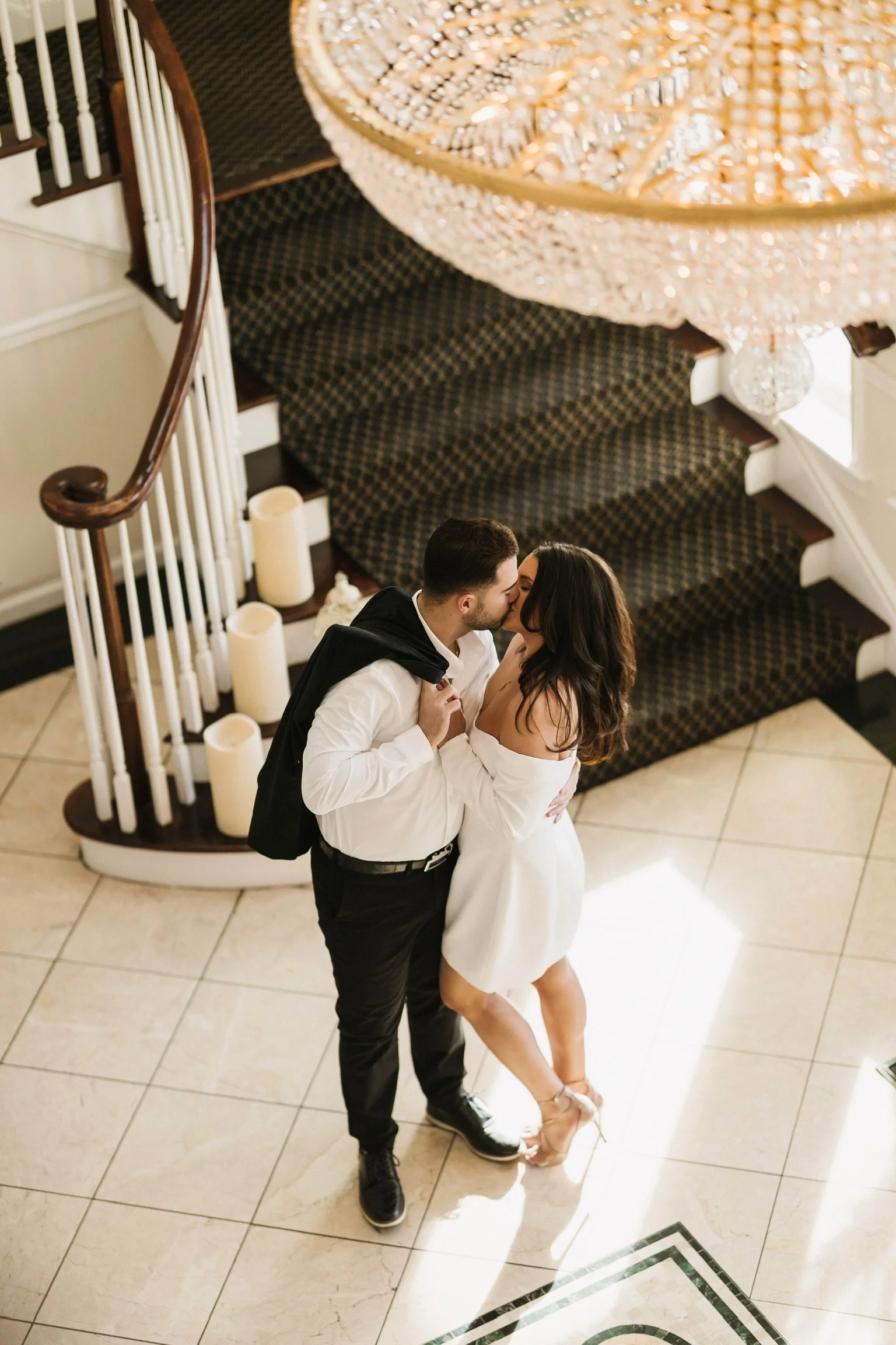 A man and woman kissing on a staircase in an indoor setting, with candles nearby and a chandelier overhead.