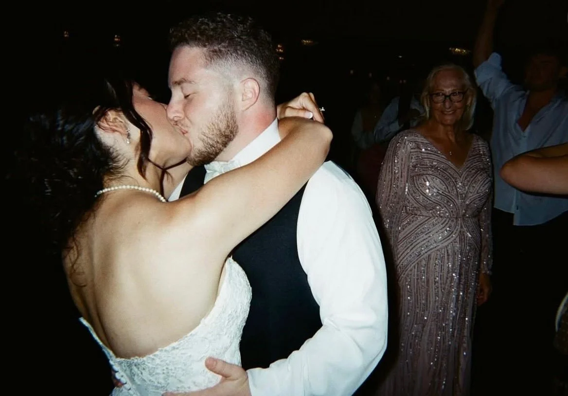 A bride and groom sharing a kiss during their wedding reception, surrounded by guests, including an older woman in a sparkly dress smiling in the background.