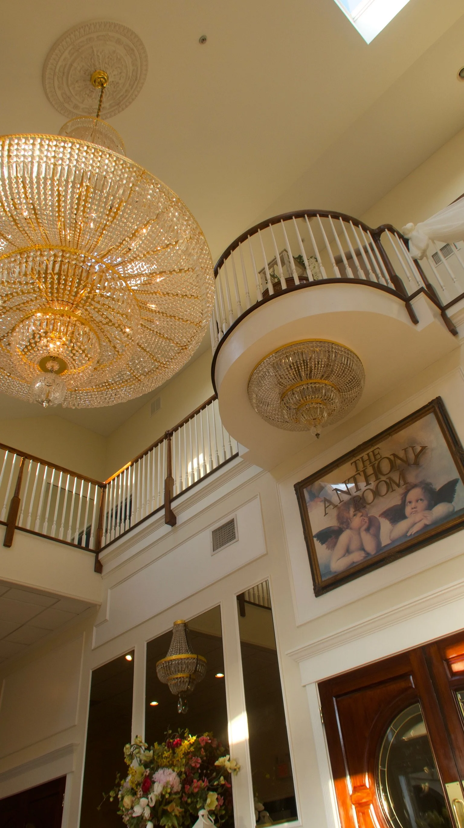Inside a grand building with ornate chandeliers hanging from the ceiling, a circular balcony with white and dark wood railings, a large framed painting titled 'The Anthony Room' featuring cherubs, and a floral arrangement underneath a chandelier.