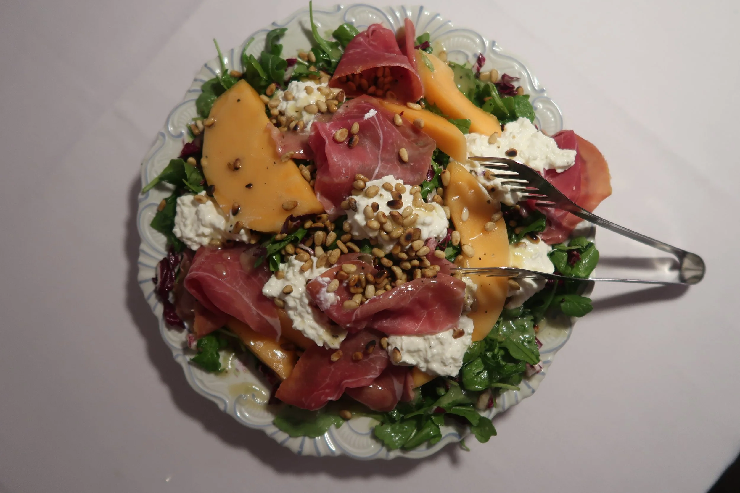 Salad with mixed greens, cantaloupe slices, prosciutto, mozzarella cheese, sunflower seeds, and a dressing, served on a decorative white plate.