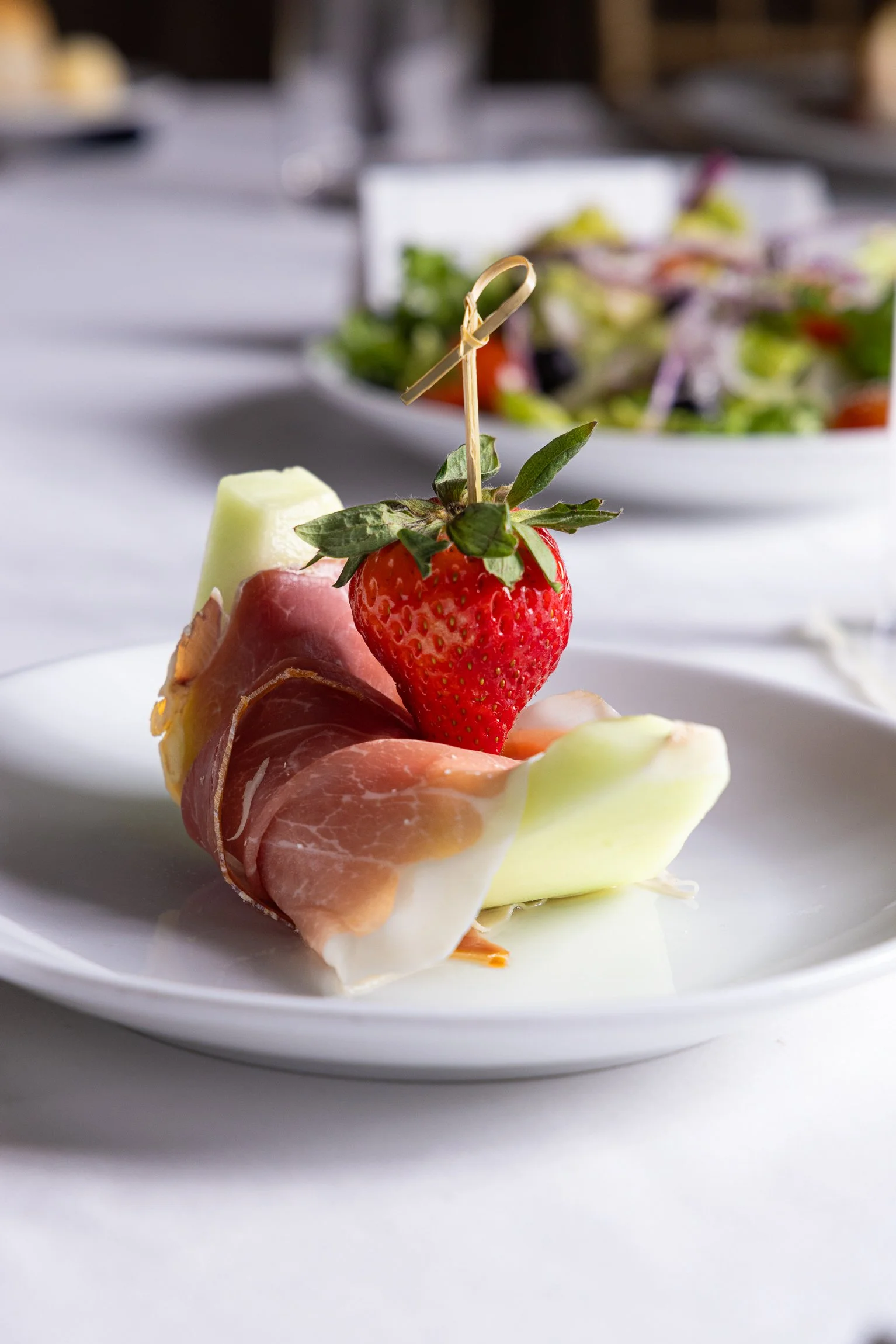 An appetizer with a strawberry on top, wrapped in prosciutto, and garnished with a small piece of melon, served on a white plate.