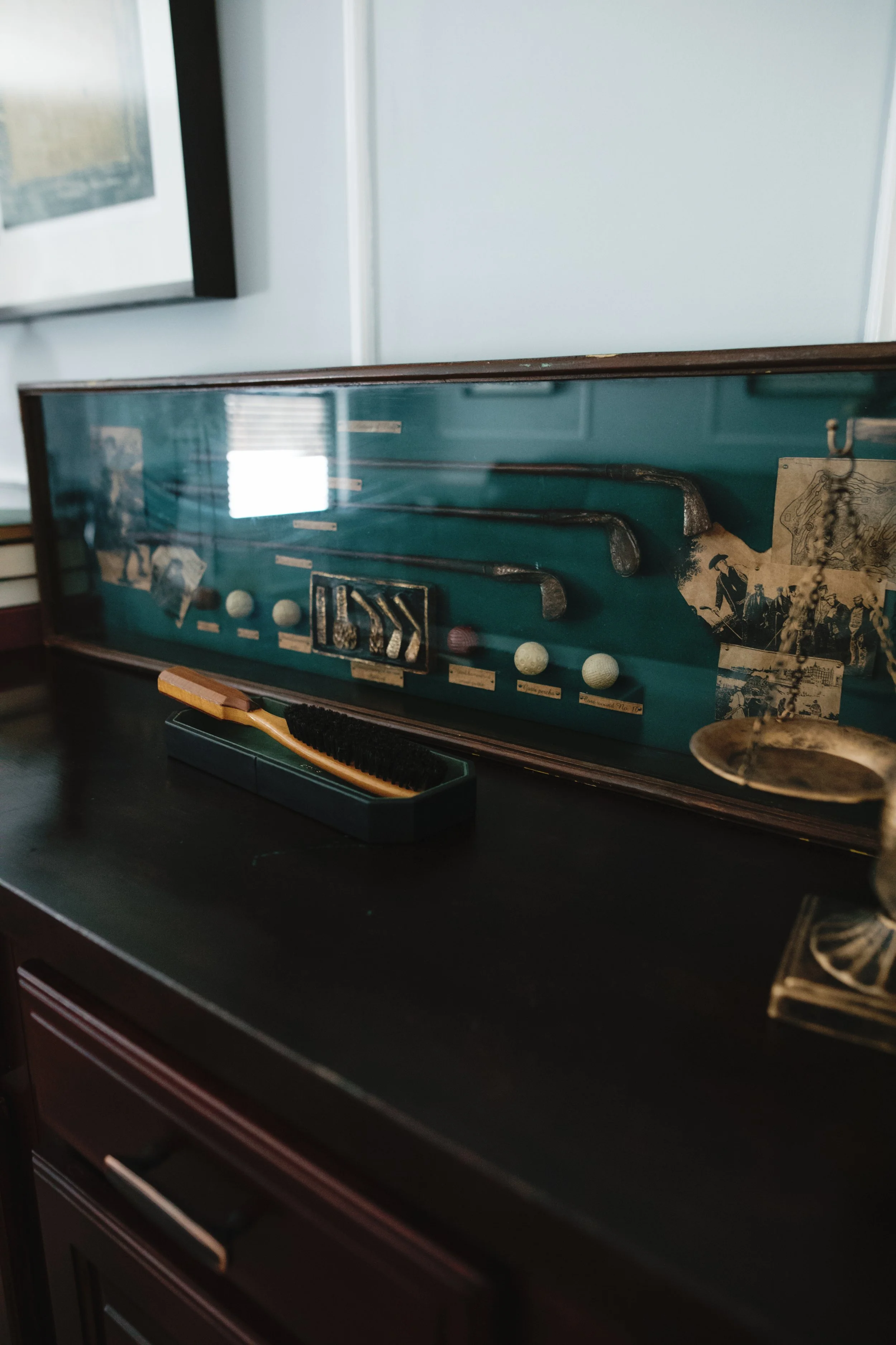 Display case with vintage golf clubs and golf balls, some old photographs, and a brush on a dark wooden surface.
